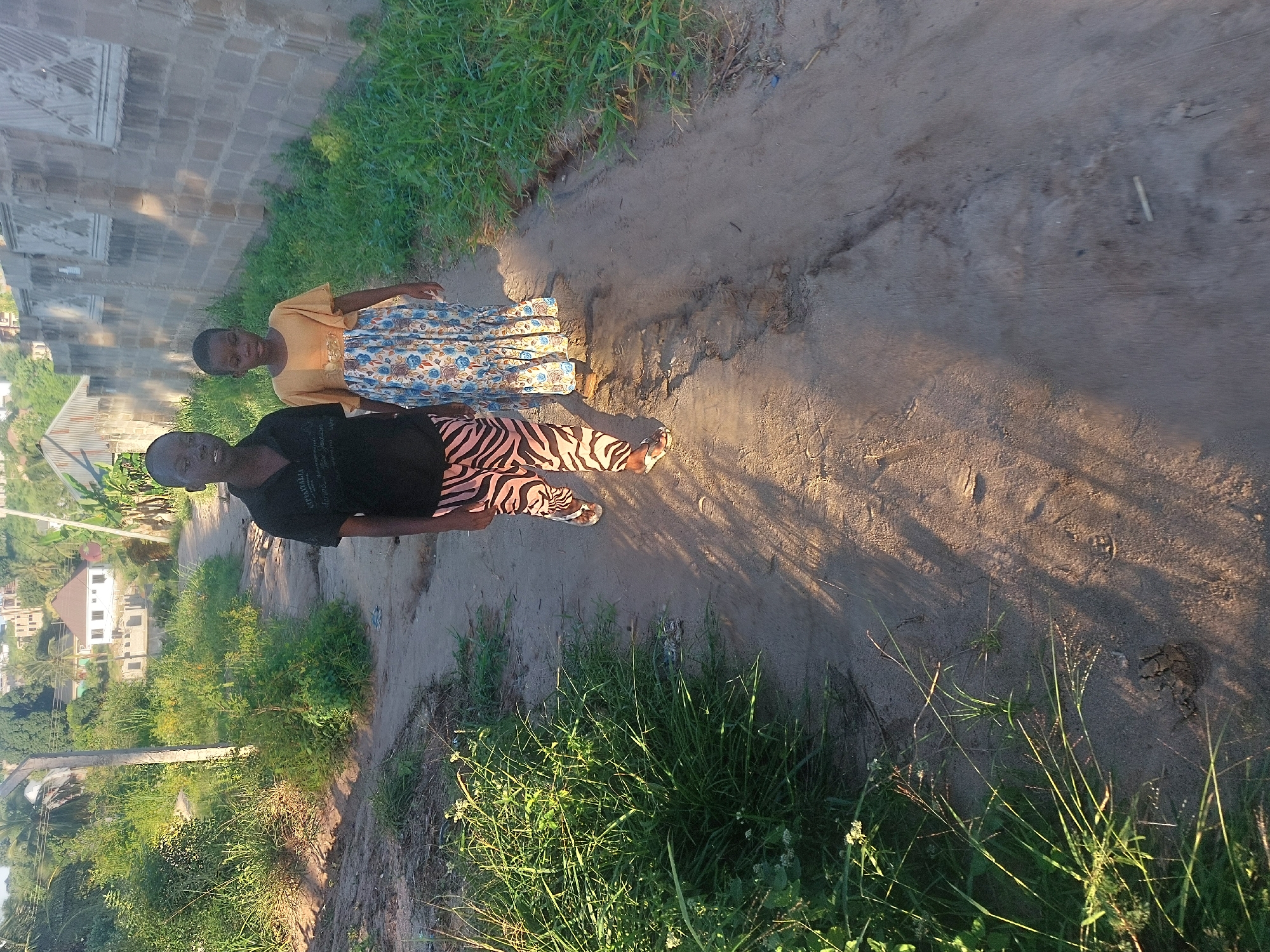 Two people stand on a dirt road. The person on the left wears a black shirt and patterned pants. The person on the right wears a yellow shirt and blue patterned skirt. The road has grass growing on either side. A building is on the left and houses are in the background. The image was taken in Dar es-Salaam, Tanzania.