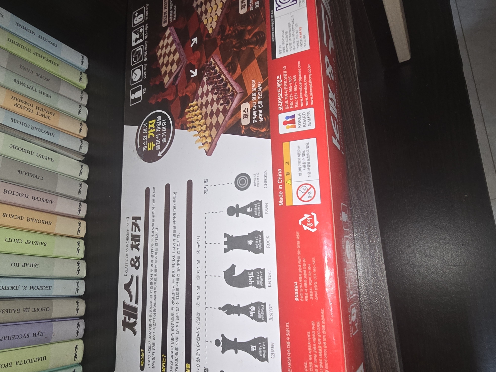 The image displays the packaging for a "Chess & Checker" board game. The packaging is primarily red and white, with Korean text and graphics. It features illustrations of chess pieces, a chessboard, and symbols related to game rules and age recommendations. A warning about small parts is also visible. The packaging is placed on a dark, wooden shelf, likely in a retail environment or a storage area. Several books with Cyrillic lettering are stacked behind the packaging, suggesting a connection to Russian literature or a location where Russian language materials are present. The overall scene is indoors, with artificial lighting. There is no identifiable information in the image that would place it specifically in the city of Ulan-Ude, Russia.