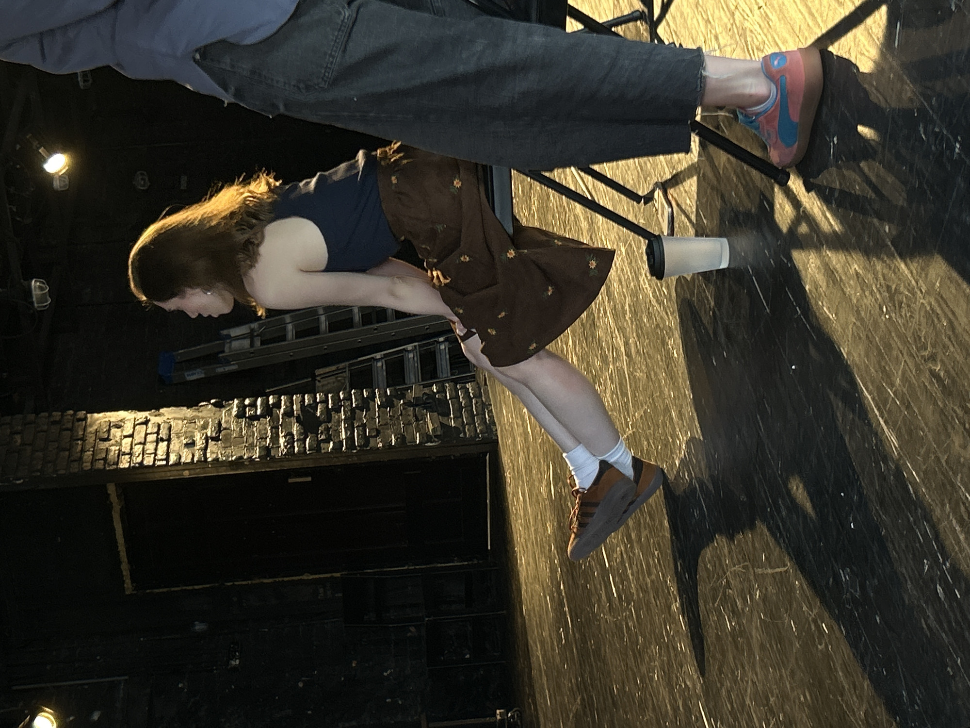 A low-angle shot shows a young woman with long hair, wearing a navy blue top and a brown floral skirt, bending over towards a brick wall and a metal structure that appears to be a ladder. She is wearing brown sneakers with white socks. Above her, a person wearing a light blue shirt and dark gray pants is seated on a black metal chair, their feet visible in the upper right corner, clad in pink and blue sneakers. A white cup or container sits on the floor beneath the chair. The scene appears to be indoors, possibly a theater or studio with dark walls and a wooden floor. The location is unknown, Unknown.