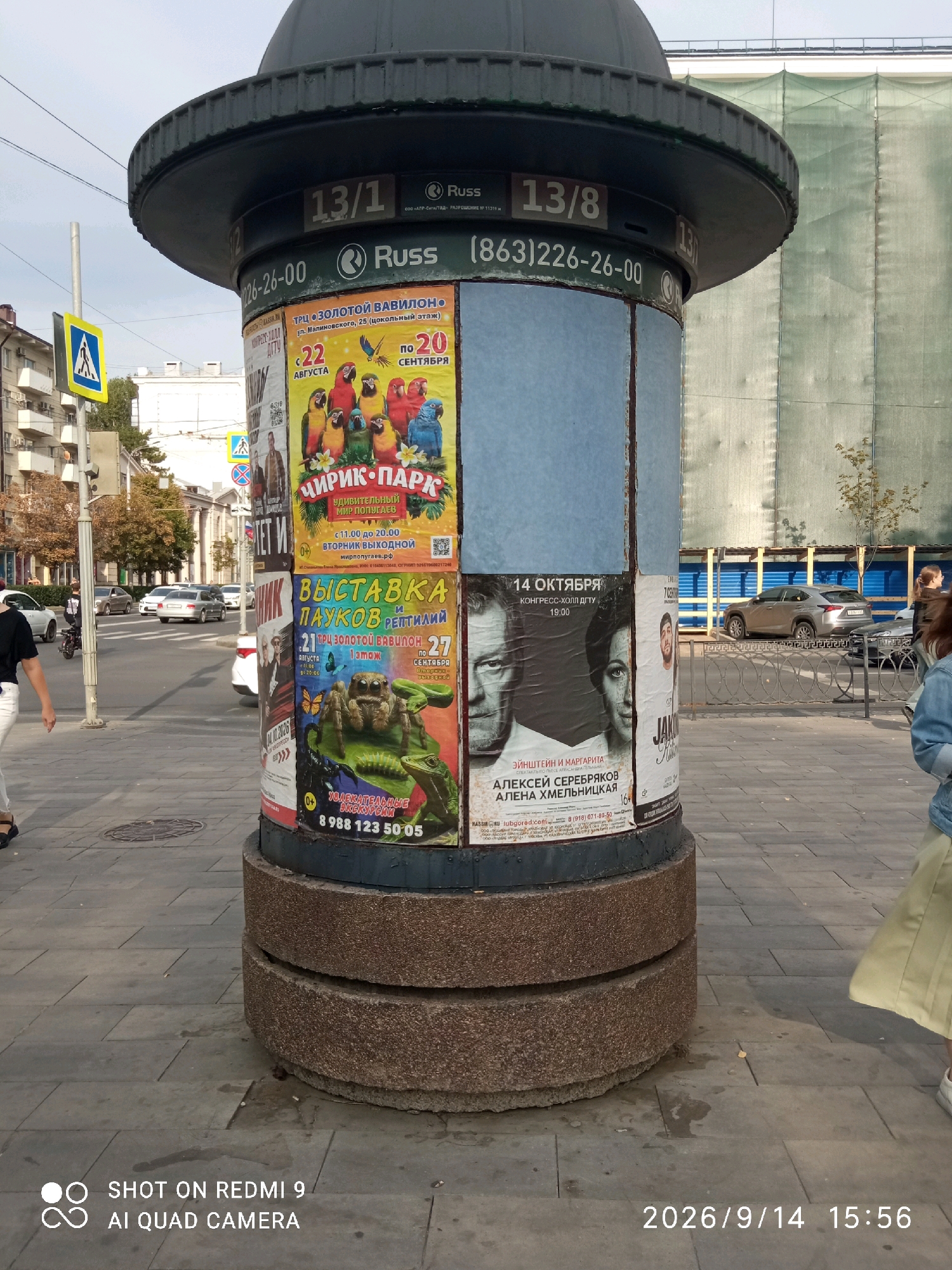 The image shows a cylindrical public advertisement pillar located on a paved sidewalk in an urban environment. The pillar is covered in various posters advertising events and attractions.
On the pillar, several posters are visible. One poster, with a yellow background, advertises "ЧИРИК-ПАРК" (Chirik-Park), which is described as "удивительный мир попугаев" (an amazing world of parrots). This poster indicates a duration from "22 августа" (August 22) to "20 сентября" (September 20) and a time from "с 11.00 до 20.00" (from 11:00 to 20:00). Another poster, predominantly blue and white, features portraits of two individuals, identified as "ЭЙНШТЕЙН И МАРГАРИТА" (Einstein and Margarita), and lists actors "АЛЕКСЕЙ СЕРЕБРЯКОВ" (Alexey Serebryakov) and "АЛЕНА ХМЕЛЬНИЦКАЯ" (Alena Khmelnitskaya). This poster also indicates a date of "14 ОКТЯБРЯ" (October 14) and a time of "19:00." Another poster, with a dark background, shows a close-up of a tarantula and a lizard, and advertises "ВЫСТАВКА ПАУКОВ И РЕПТИЛИЙ" (Exhibition of Spiders and Reptiles) at "ТРЦ ЗОЛОТОЙ ВАВИЛОН" (TRC Golden Babylon) on August 21-27. A phone number "8 988 123 50 05" is visible on this poster.
Above the posters, the pillar has horizontal bands with numbers and text. One band shows "13/1" and "13/8". Below this, there is text that appears to include a phone number "(863)226-26-00".
In the background, a street with traffic is visible, including cars and a person on a motorcycle. Pedestrians are present on the sidewalk. Buildings line the street, and a large green mesh structure, likely under construction or renovation, is prominent in the background. A pedestrian crossing sign with a yellow background and a blue symbol is also visible. The ground is paved with grey square tiles.
The bottom right corner of the image contains a watermark indicating "SHOT ON REDMI 9 AI QUAD CAMERA" and a date and time stamp "2026/9/14 15:56".
Based on the phone number format "(863)", which is a common area code for Rostov-on-Don, and the Cyrillic text on the advertisements, the location is likely Rostov-on-Don, Russia.