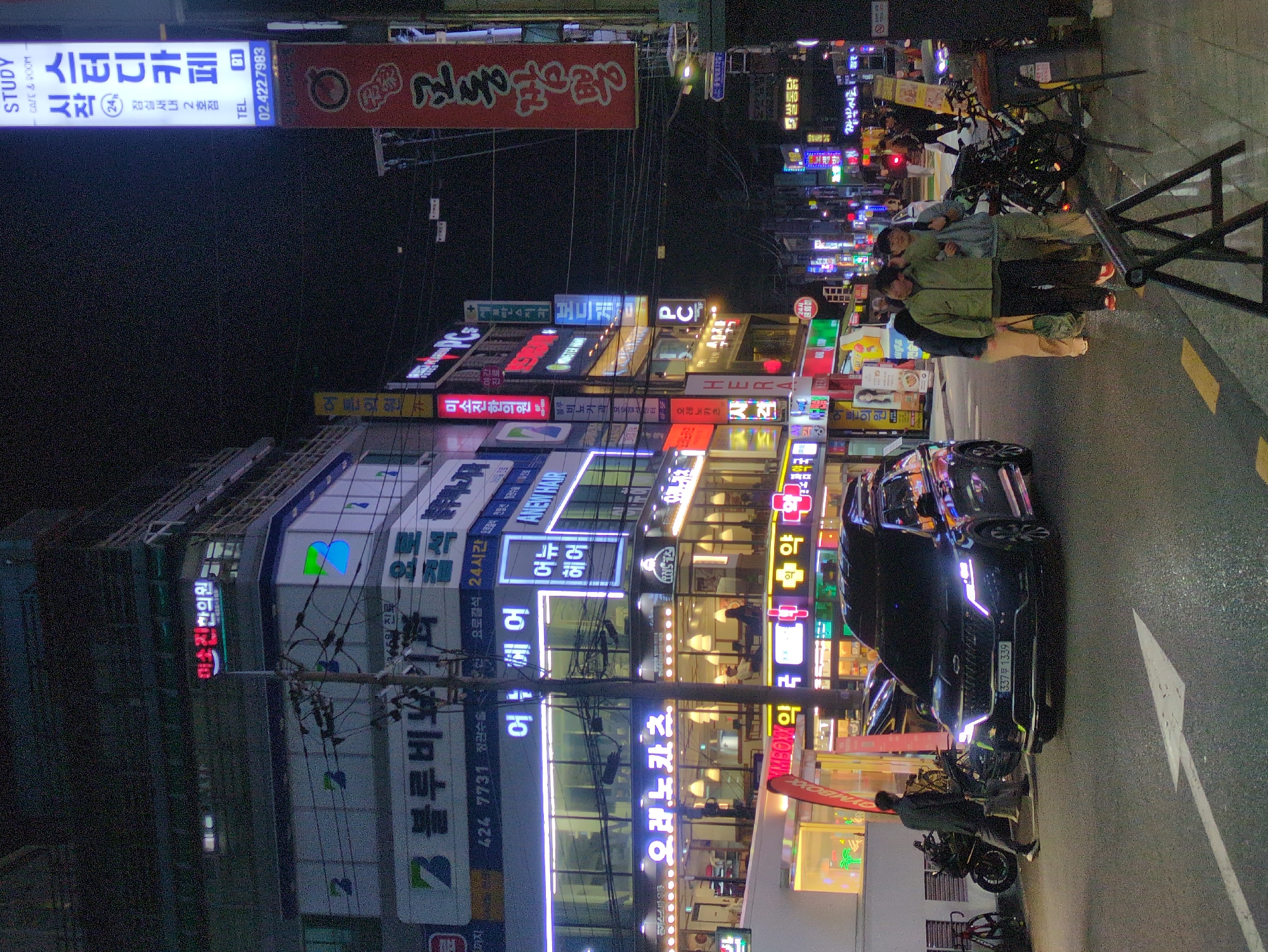The image depicts a street scene at night in Seoul, South Korea. The scene is dominated by brightly lit buildings with multiple Korean-language signs. Streetlights cast a glow on the wet asphalt road. A black car is parked on the right side of the road, partially obstructing a motorcycle. An individual is interacting with the motorcycle, seemingly parked near the sidewalk. Several pedestrians are standing near a metallic barrier. The vertical orientation of the image makes it difficult to determine perspective. The street appears to be active with commercial activity. The intersection is marked with a white arrow on the road surface.