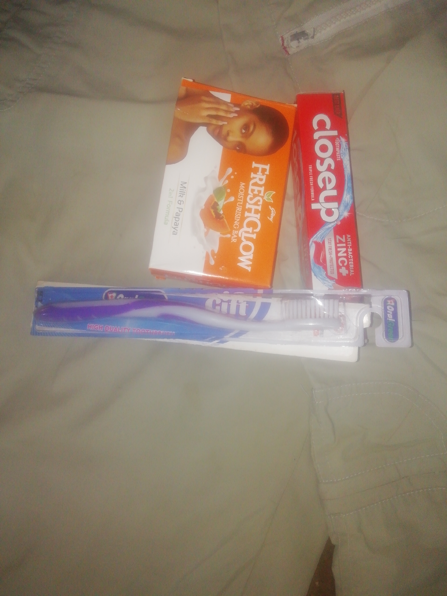 The image displays a flat lay of personal hygiene products against a light-colored, possibly beige or khaki, fabric surface that appears to be clothing, indicated by a visible zipper and stitching in the upper right. There is no discernible activity taking place.
The main subjects are three packaged items. On the left, an orange box for "FRESHGLOW MOISTURISING BAR" by "Godrej" is visible, with text indicating "Milk & Papaya" and "2in1 Formula." The box features an image of a woman's face and fruit. To its right, a red box of "closeup TOOTHPASTE" is shown, bearing the descriptions "TRIPLE FRESH FORMULA," "ANTI-BACTERIAL ZINC+," and "STAY FRESH PROTECTED." Below these, a package containing two toothbrushes is laid horizontally. The brand name appears to be "cifft" or similar, with the text "HIGH QUALITY TOOTHBRUSH" clearly visible. One toothbrush is purple and white, the other is white. A small logo on the packaging might read "Oral Greet" or similar, suggesting "Oral-B." The lighting is subdued, and the image is somewhat blurry.
