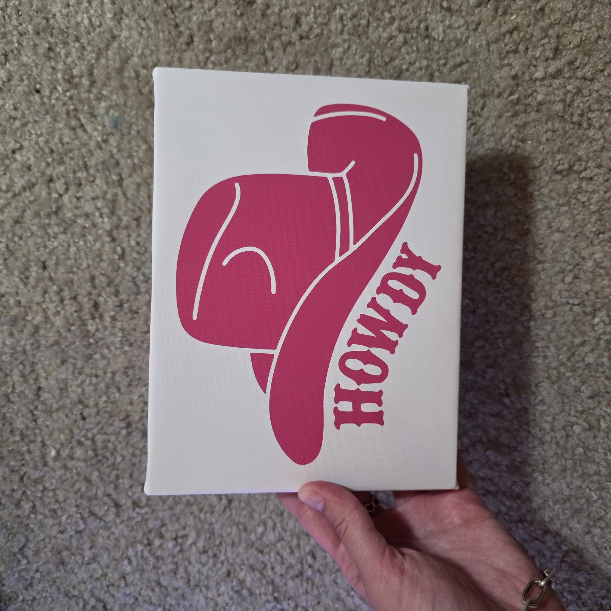 A hand is holding a small, rectangular canvas art piece. The canvas is white and features a large, hot pink graphic of a cowboy hat. Below the hat, the word "HOWDY" is written in the same hot pink color, in a decorative, Western-style font. The canvas is placed on a beige carpet. The hand holding the canvas has a silver bracelet on the wrist and a ring on one finger.