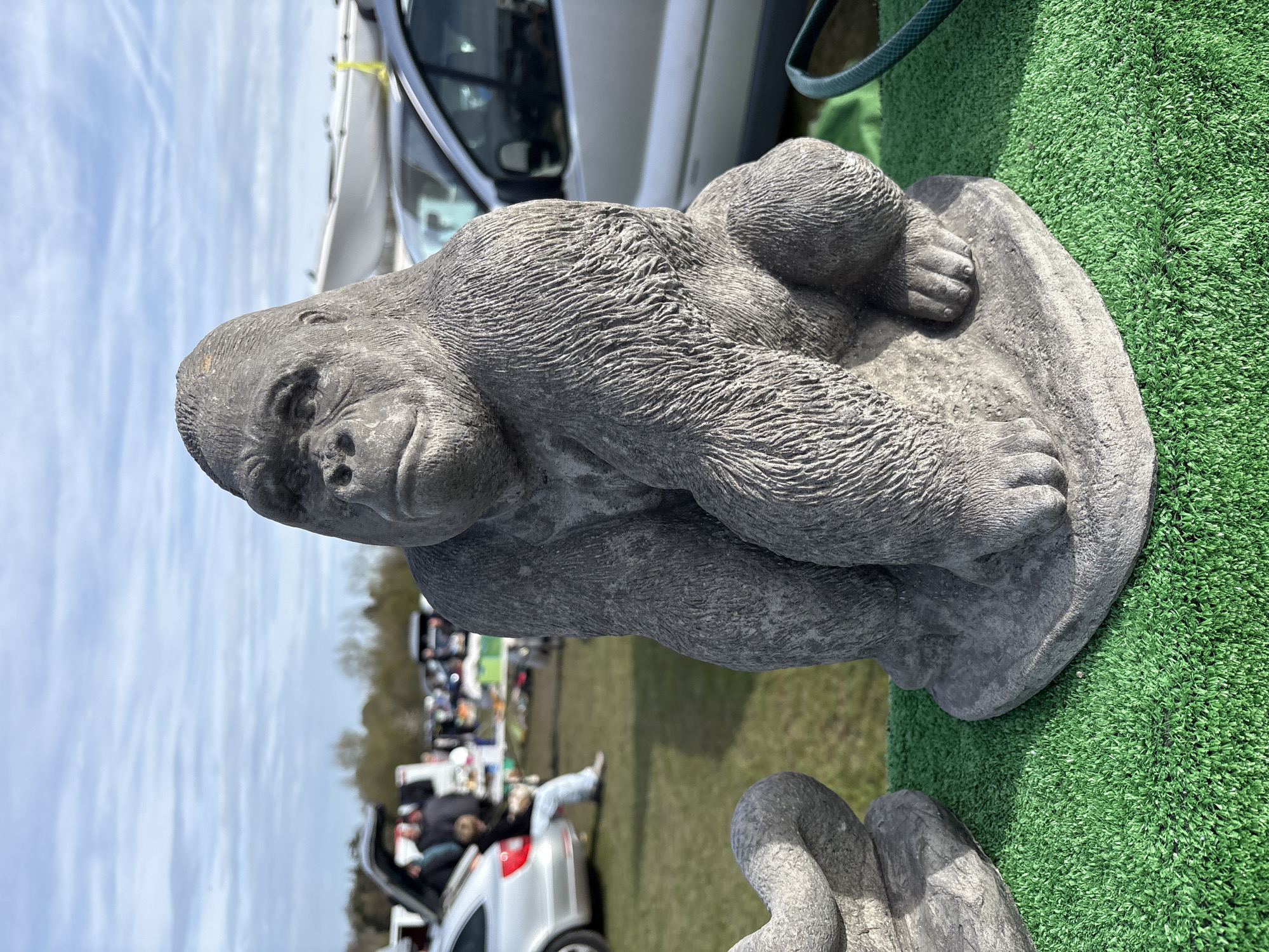 A concrete gorilla statue sits on green artificial turf. A car and people are visible in the background, along with other vehicles and objects suggesting a flea market or outdoor sale. The location appears to be outdoors, possibly in a grassy area within the city of Halton, United Kingdom.