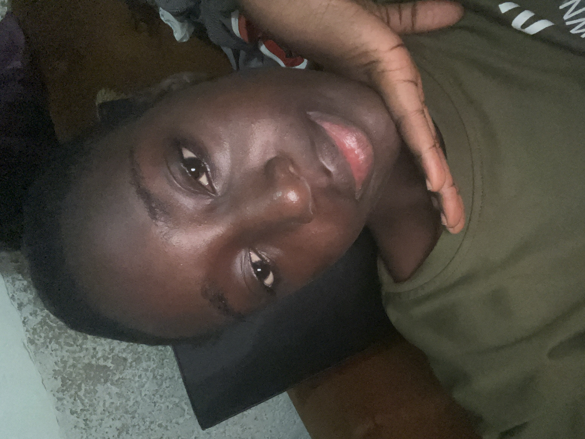 A close-up, angled shot depicts a Black person wearing an olive green shirt, lying down. The person’s face is prominently visible, with their eyes open. A hand is positioned near their face. The background appears to be a combination of flooring and what looks like clothing. There is no contextual information to suggest this photo was taken in Abuja, Nigeria.
