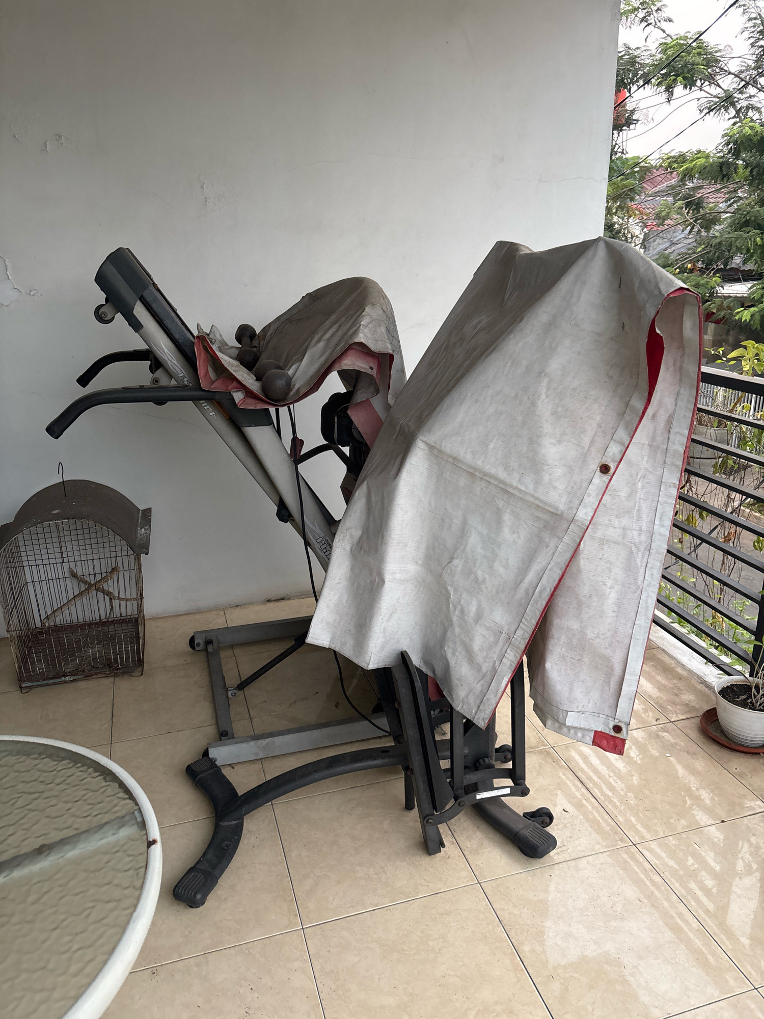 The image depicts a covered exercise machine, possibly a treadmill, and a birdcage on a tiled outdoor patio. The exercise machine is partially covered by a grey tarp with red trim. Several dark, spherical objects, likely dumbbells, are visible beneath the tarp. To the left of the exercise machine, an old, metal birdcage with a wooden perch stands. In the foreground, a portion of a round glass-topped table with a white rim is visible. The patio is paved with light-colored square tiles, some of which appear wet. In the background, a white wall, a railing, and lush green trees with distant rooftops can be seen. The scene suggests a residential outdoor space in Bekasi, Indonesia, possibly a balcony or patio.