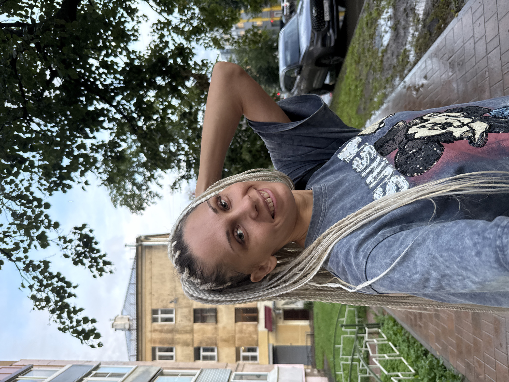 The image shows a woman with blonde braided hair posing for a selfie outdoors. She is wearing a dark gray t-shirt with a sequined graphic of Mickey Mouse on the front. Her left arm is raised, and she is touching the back of her head. She is smiling at the camera. The background consists of a residential building with yellow and gray facades, a tree with green leaves, a wet sidewalk, a patch of grass, and a parked car. The location is identified as Fedorino, Russia.