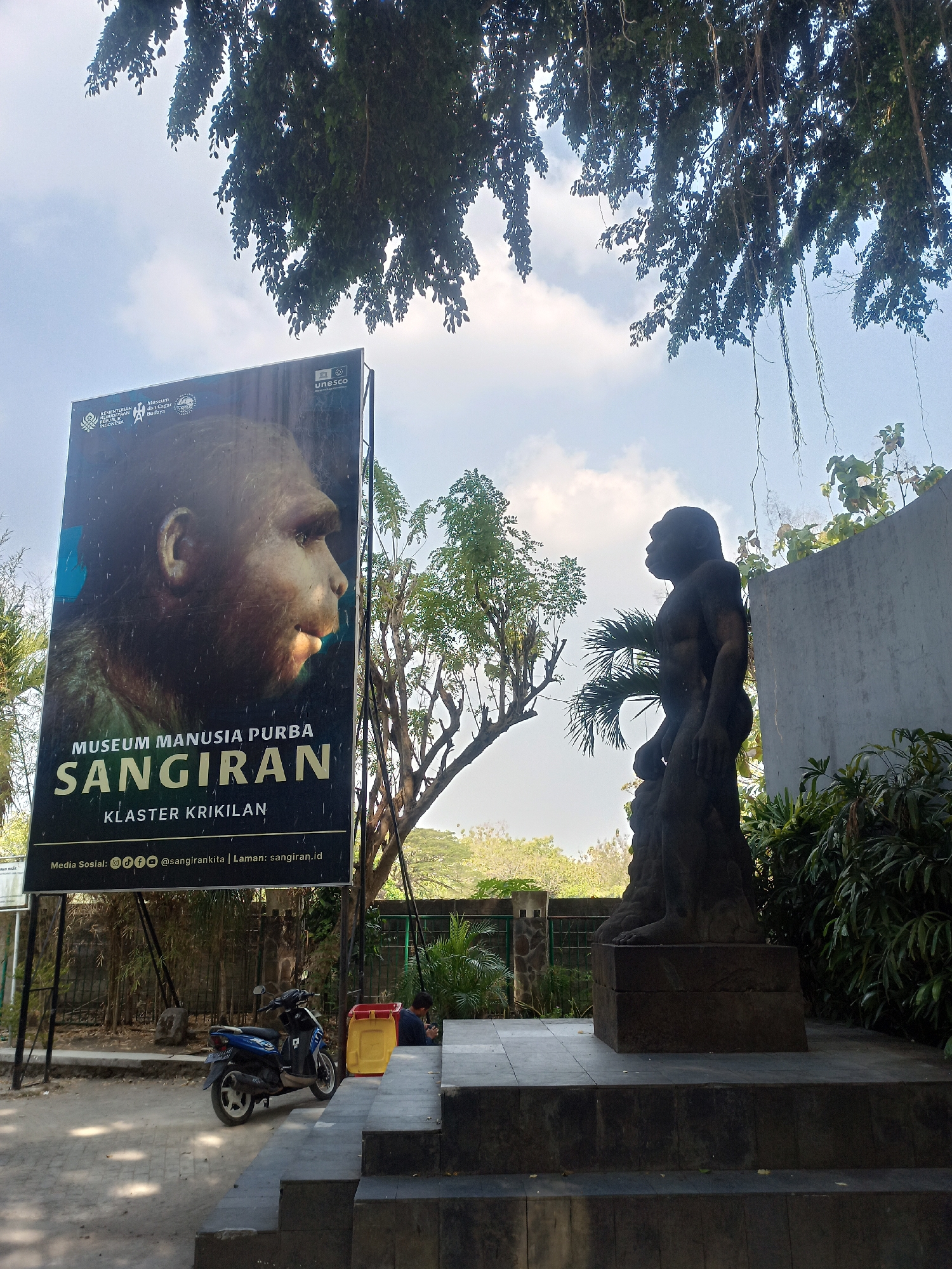 The image depicts the entrance to the Sangiran Early Man Museum in Gondangrejo, Indonesia. A large billboard prominently displays an image of an early human's head with the text "MUSEUM MANUSIA PURBA SANGIRAN" and "KLASTER KRIKILLAN". To the right of the billboard, a dark, stone statue of an upright-standing hominid is positioned on a tiered platform. In the background, lush green foliage and trees frame the scene, with a bright sky visible above. In the mid-ground, a blue and black scooter is parked on a dirt path. A person is seated on the steps to the right of the scooter, near a yellow and red trash bin. The environment appears to be an outdoor museum or park setting.