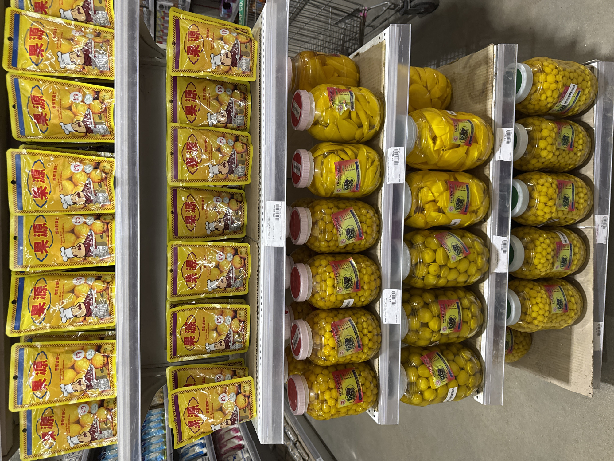 The image shows shelves in a supermarket, likely in Thong Sala, Thailand, stocked with various preserved and packaged foods. On the left, multiple packets of "Country Roasted Chestnuts" are arranged. Moving to the right, shelves contain large jars of preserved fruits or vegetables, with bright yellow contents. The jars are labeled in Thai and English. A shopping cart is visible in the background, indicating a retail environment. The shelves appear well-stocked and organized. Price tags are attached to the shelving units.