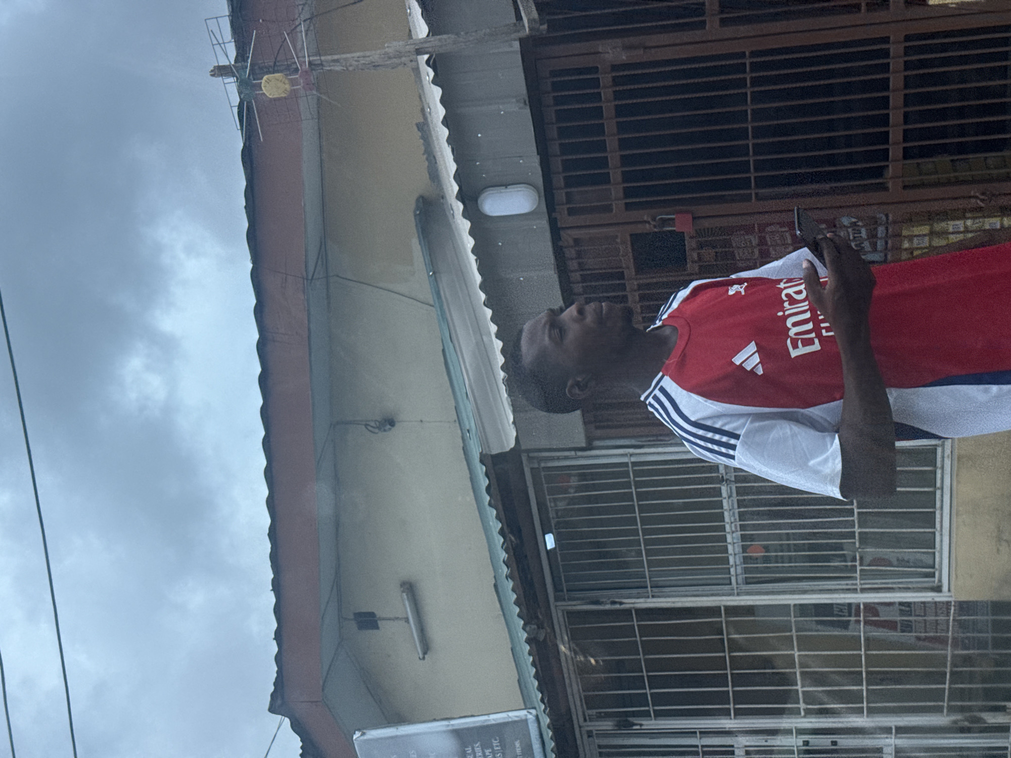 A man wearing an Arsenal jersey holds a cell phone while standing in front of a building in Ado, Nigeria. The building has barred windows and is partially obscured by an overhanging roof. Overhead cables and a cloudy sky are visible.
