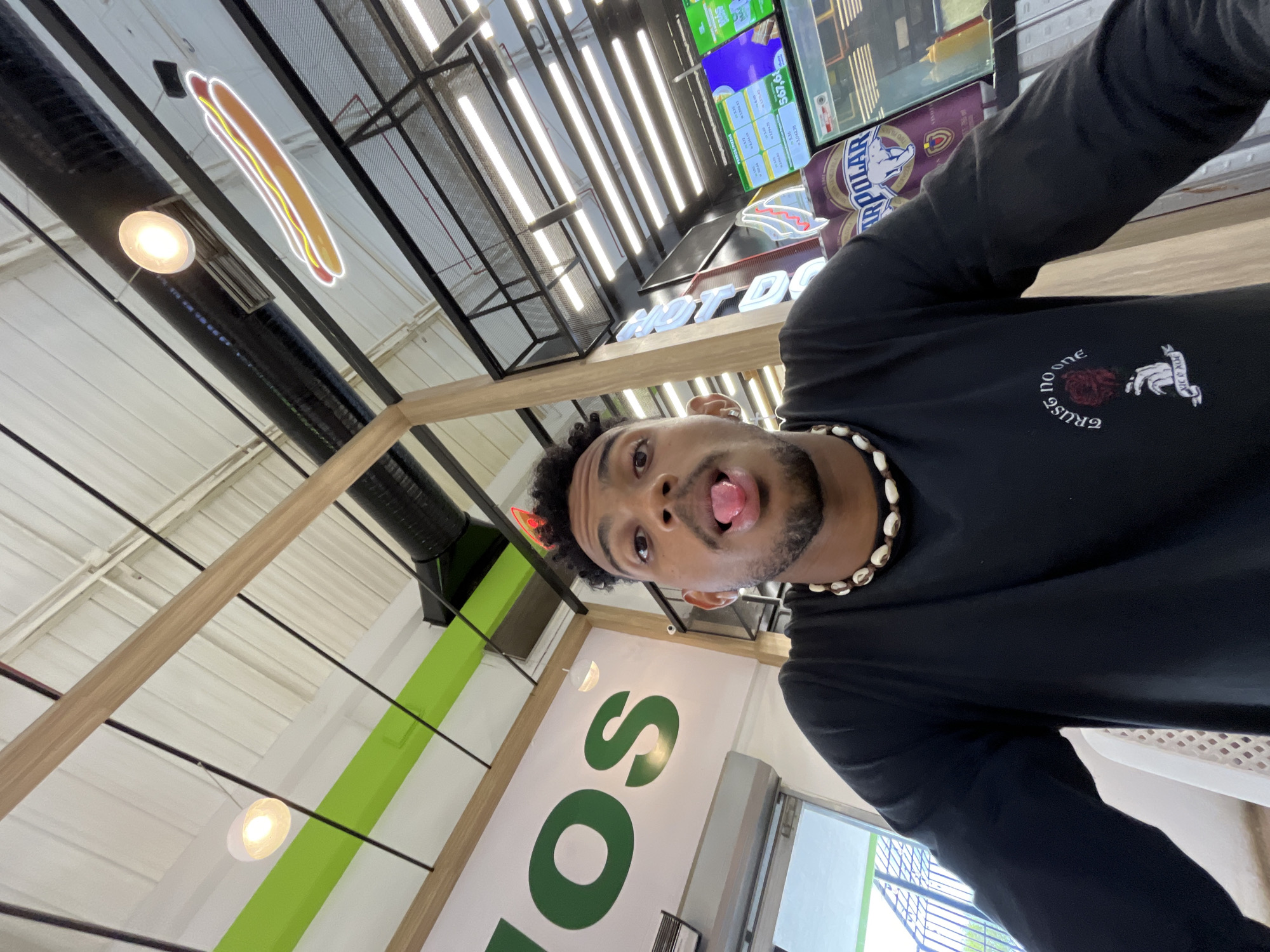 The image presents a low-angle selfie of a single male subject positioned centrally, his tongue extended. The indoor setting is identified as being in Porlamar, Venezuela, based on the visible "ARPOLAR" branded cans (a Venezuelan beer) and digital menu screens displaying "PEDIDOS/PIZZAS" with prices denoted in Bolívares (e.g., Bs. S 1.043.75). The subject is dark-skinned, with dark curly hair and a beard, and wears a black long-sleeved shirt featuring a white embroidered design on the left chest: "TRUST NO ONE" above a red rose held by a skeletal hand, with "XOX & XOX" below. He is also wearing a necklace made of irregular white shell beads. The background includes a wall with large green letters spelling "OS", suggesting part of a larger sign. The ceiling consists of white corrugated metal panels, exposed black ventilation ducts, wooden beams, and black metal structural elements. Lighting is provided by spherical pendant lights and recessed strip lights. A neon hot dog sign is suspended from the ceiling. A partially visible white plastic chair and an exterior view through a window or doorway are discernible on the far left.