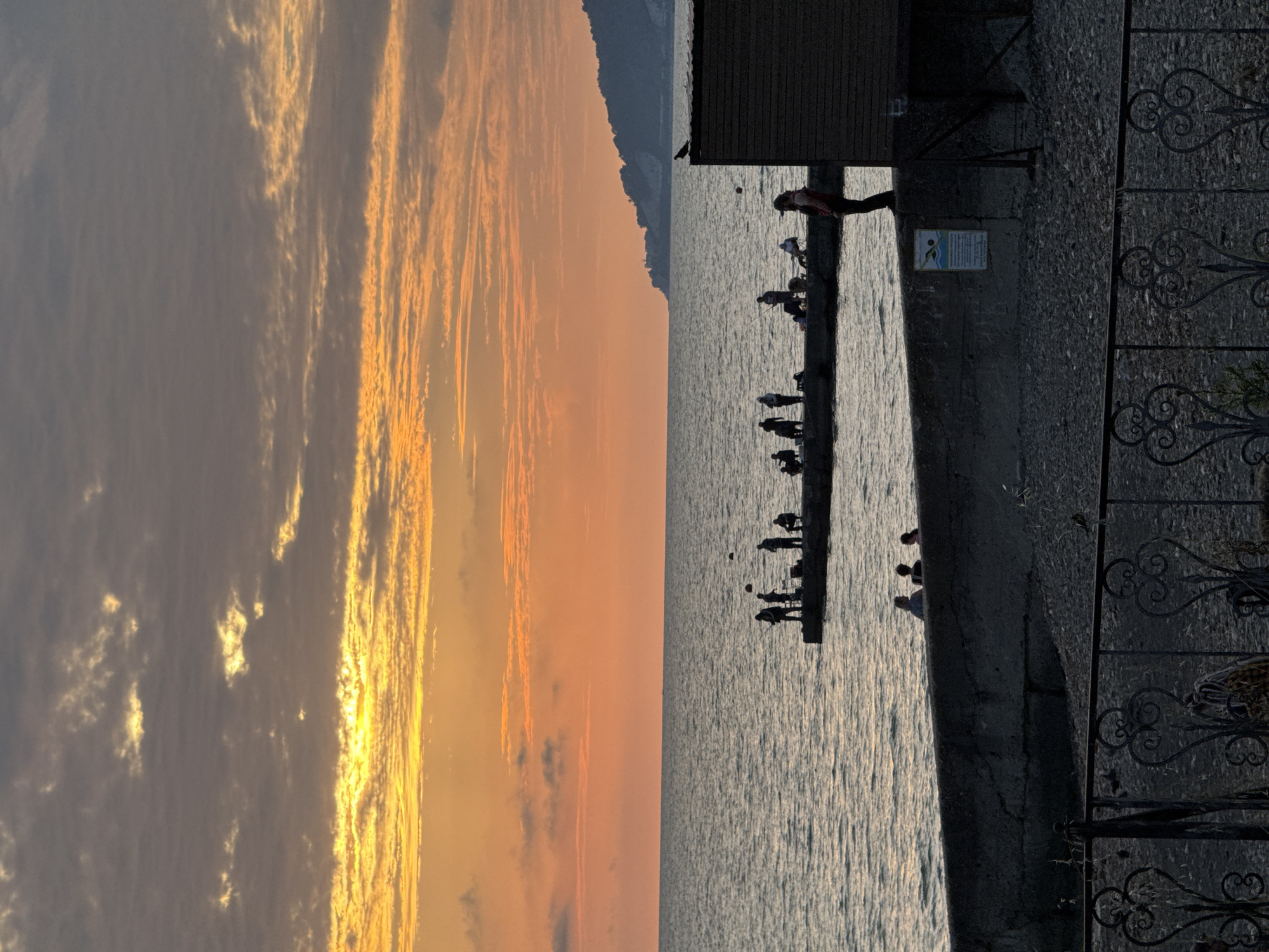 This image depicts a sunset scene on the Black Sea coast in Sochi, Russia. In the foreground, a concrete embankment lines a pebbled beach, with a decorative metal fence partially visible. Further out, a pier juts into the sea, occupied by several silhouetted figures fishing. The water is calm with gentle ripples. The sky is a gradient of orange and yellow, with scattered clouds, indicating the setting sun. A small, dark building with a sloped roof stands on the right side of the image, partially obscuring a distant, tree-covered hillside. A solitary bird is perched on the roof of the building. The overall atmosphere is peaceful and serene, capturing a moment of evening leisure on the coast.