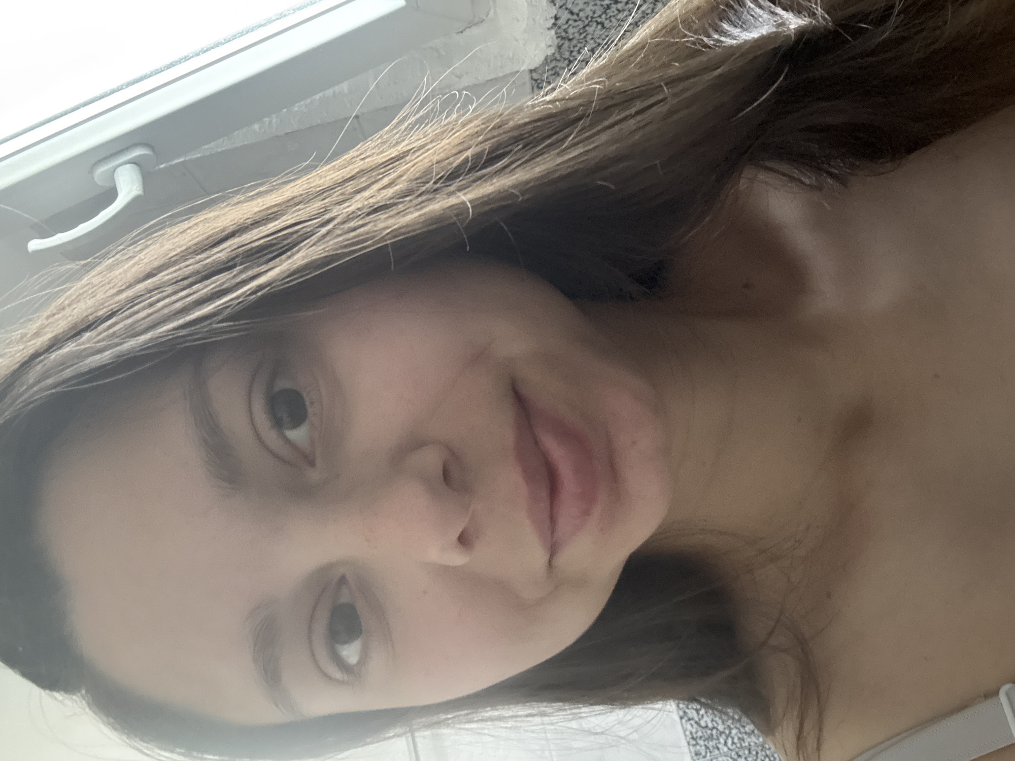 The image is a close-up, waist-up, selfie-style photograph of a young woman with long, brown hair. She is looking directly at the camera with a slight smile. Her skin is fair, and she has brown eyes. She is wearing a light-colored top. The background appears to be indoors, near a window with a white handle. The lighting suggests it is daytime. The image does not contain any elements that would allow for identification of the location as Plovdiv, Bulgaria.