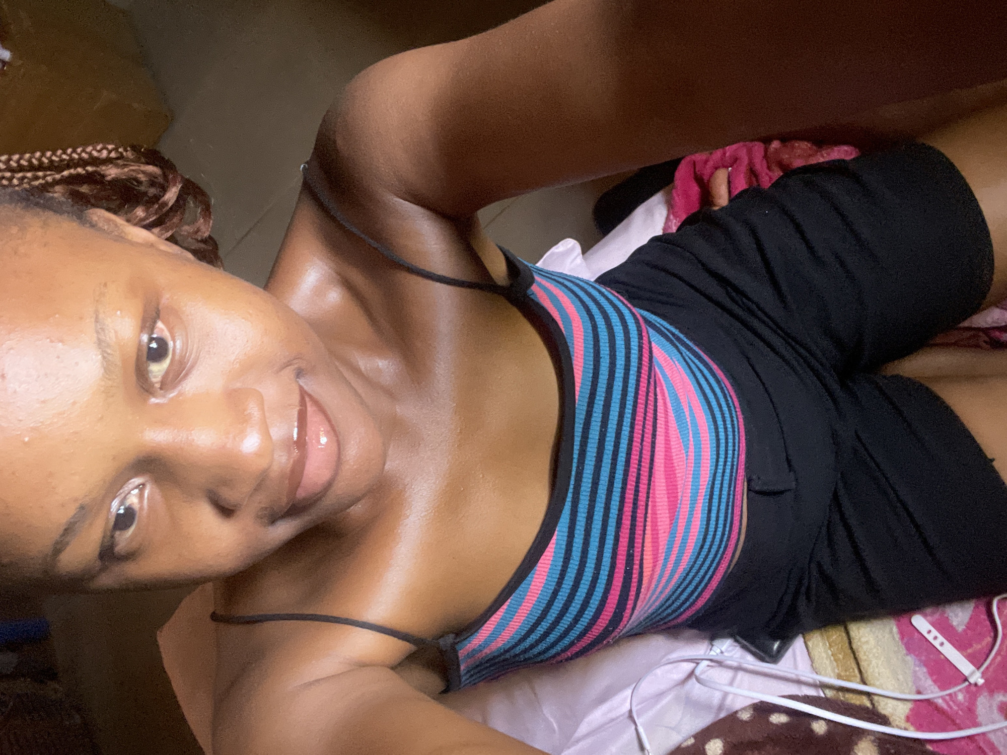 The media file is a portrait-style, self-taken photograph of a young Black woman lying down. She has her hair in braids and is wearing a striped tank top with blue and pink stripes and black shorts. The background is dimly lit, suggesting an indoor setting, possibly a bedroom, with a glimpse of patterned fabric and some pink material. A pair of white earbuds is also visible in the lower right corner. The woman's expression is relaxed and she appears to be looking at the camera. The lighting and her glossy skin suggest a warm environment, and the overall mood is casual and personal. No visible text is present in the image. The location context is Jos, Nigeria.