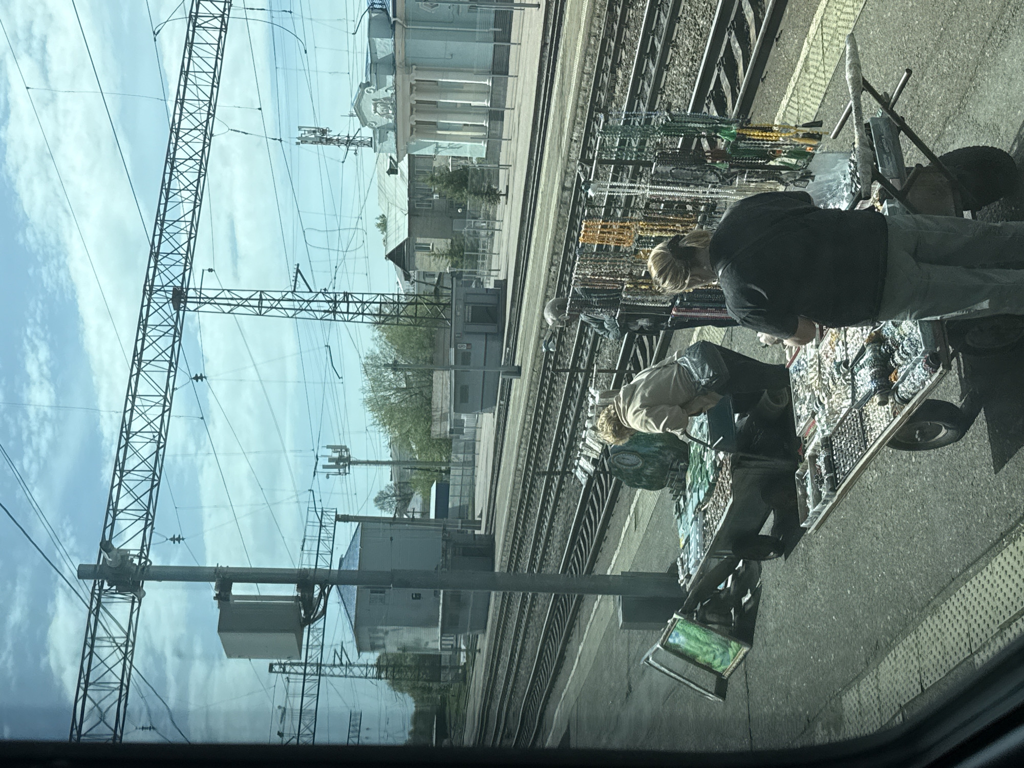 The image depicts a scene at a railway station in Kropachyovo, Russia, as viewed from inside a train. The view is across the railway tracks. There are at least two individuals near a makeshift jewelry stand or cart. The cart holds a variety of jewelry items. The background consists of railway infrastructure, including tracks, electrical lines, support structures, and buildings typical of a railway environment. The weather appears to be sunny with some clouds in the sky.