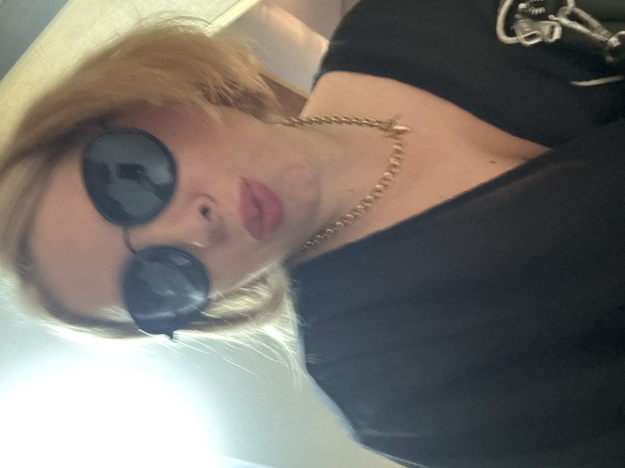 A woman with blonde hair and pink lipstick is wearing sunglasses and a black top. She is also wearing a gold chain necklace with a heart-shaped pendant. The image is a selfie taken indoors, with soft lighting. It is not possible to determine the location in the city of Sochi, Russia, from the provided image.