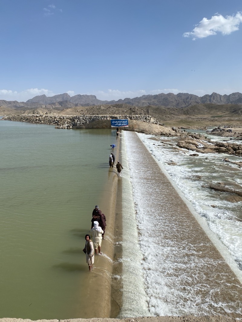 The image shows a wide shot of a weir or dam in Kim, Afghanistan. The water behind the dam is calm and pale green, while the water flowing over the dam is frothy and white. A group of men, dressed in traditional Afghan clothing, are wading across the dam. The background features arid, rocky terrain and mountains under a bright blue sky with some clouds. A sign is visible on the far side of the dam, but the text is unreadable.