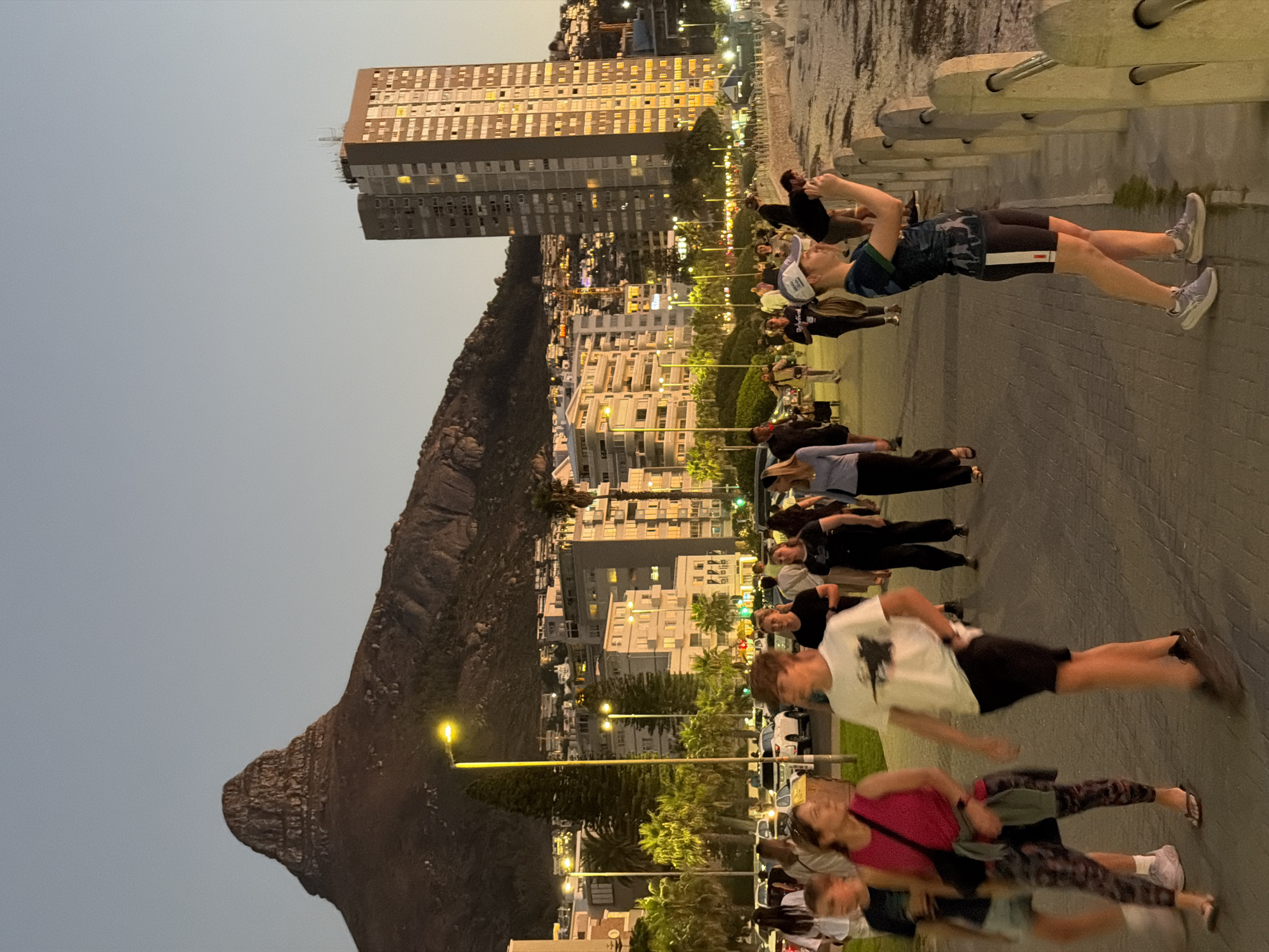 The image shows a street scene at night in Cape Town, South Africa. In the background is Lion's Head mountain and several buildings, including a tall apartment building. A crowd of people is walking or standing along a paved promenade next to a low wall, possibly a sea wall. Street lights illuminate the scene. People are of various ages and are dressed casually. Some are walking quickly, resulting in motion blur. Some individuals near the wall are looking toward the water.