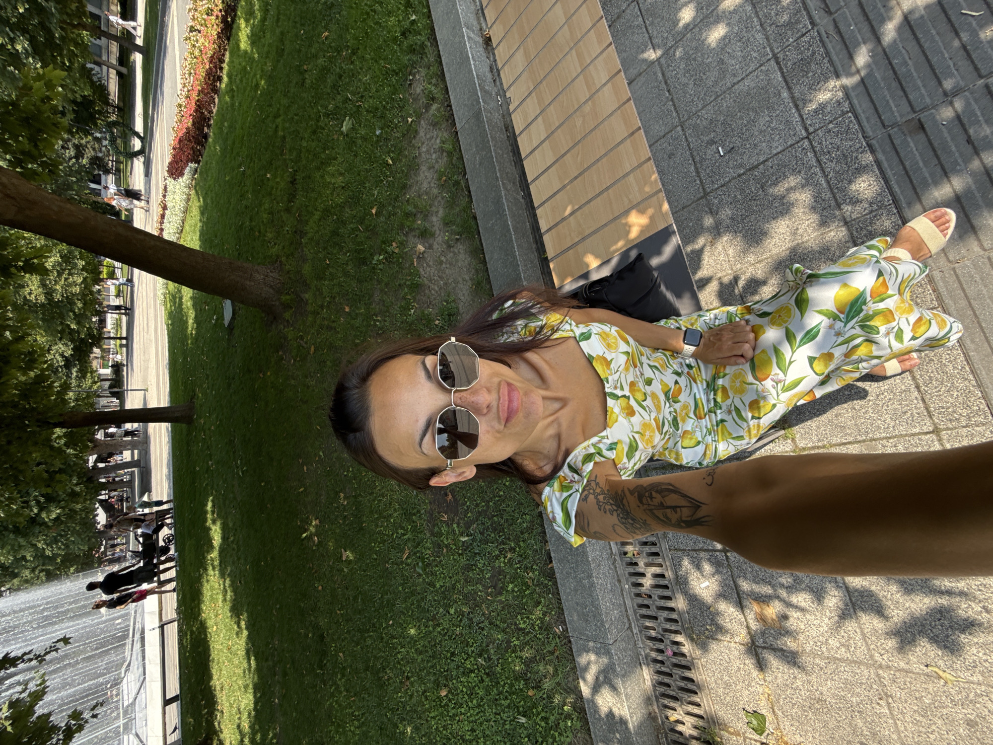 In a selfie taken outdoors on a sunny day, a woman with long dark hair and sunglasses is seated on a bench. She is wearing a white dress with a yellow lemon print and beige sandals. Her left arm, which is tattooed, is extended towards the camera, holding the phone for the picture. She has a slight smile on her face.
The background shows a park setting with lush green grass, trees, and a flowerbed with red and pink flowers. Further in the distance, there is a fountain with water spraying, and several people can be seen walking along a path. The ground is paved with grey tiles and some wooden planks form part of the bench. The lighting suggests it is daytime, likely late morning or afternoon, with dappled sunlight filtering through the trees. The overall mood appears relaxed and casual.