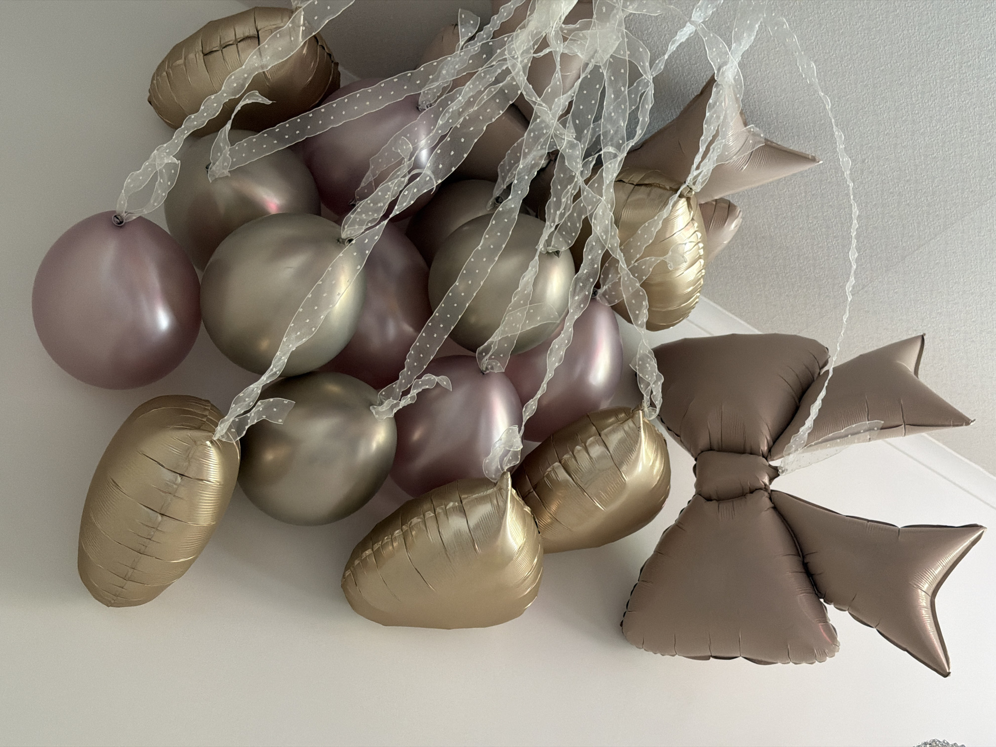 The image presents a cluster of balloons suspended against a plain white ceiling, indicating an indoor setting decorated for an event. The balloons display a palette of muted metallic and pastel shades, predominantly rose gold/lilac, silver-grey, and gold, along with a brownish-grey.
The collection includes a mix of balloon types. Some are round, seemingly latex balloons, in shades of rose gold/lilac and silver-grey, appearing fully inflated and smooth. Interspersed among these are various foil balloons. Several gold foil balloons, some appearing heart-shaped, show visible creases, suggesting they may not be fully inflated or have been stored. A large, prominent foil balloon shaped like a decorative bow, in a muted brownish-grey color, is situated on the right side of the frame, also exhibiting creases on its surface.
All balloons are tied with sheer white ribbon, which is patterned with small white polka dots. The ribbons extend downwards and intertwine, contributing to the decorative arrangement. The overall impression is one of a festive or celebratory atmosphere. There is no visible text within the image. While the location is contextualized as Moscow, Russia, this detail is not visually verifiable from the image content itself.