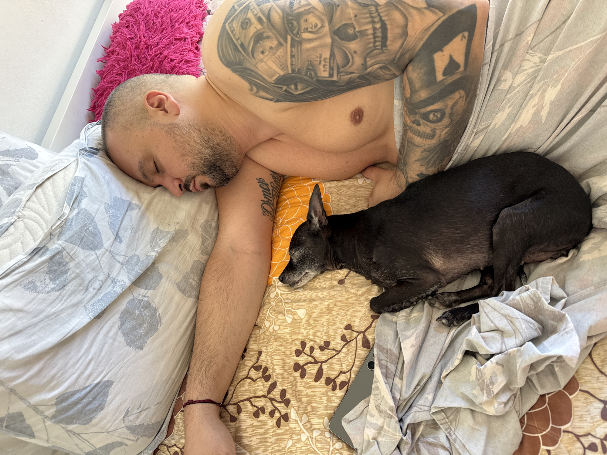 The image shows a man and a dog sleeping in a bed. The man is lying on his back with his head on a pillow. He has a beard and tattoos on his arms. The dog is black and is lying next to the man, with its head on the man's arm. The bed is covered with a floral patterned sheet. The room appears to be a bedroom, with a white headboard visible in the background. There is also a pink fuzzy pillow on the bed. The image does not provide any information about the specific location in the city of Plovdiv, Bulgaria.