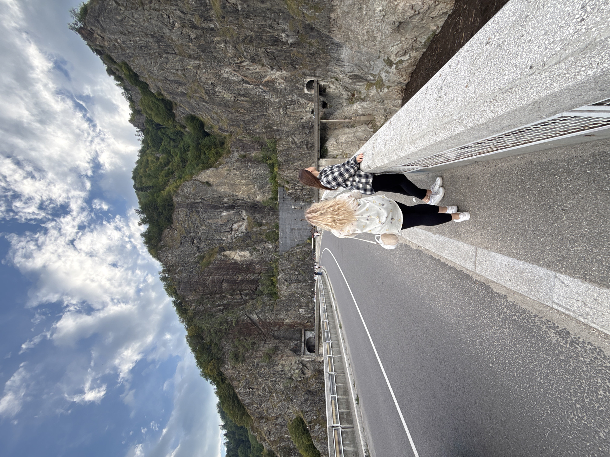 The image captures a scenic view from the Vidraru Dam in Arefu, Romania. Two young women are standing on the dam's pedestrian walkway, looking out at the dramatic mountainous landscape. The woman on the left has long, blonde, wavy hair and is wearing a white, floral-patterned shirt and black leggings. She has a small, round, beige purse slung over her shoulder. The woman on the right has long, dark brown hair and is wearing a black and white checkered shirt and black leggings. Both women are wearing white sneakers.
The dam itself is a prominent feature, with a wide asphalt road curving to the left, marked by a white dividing line. A concrete barrier with a metal railing runs along the edge of the walkway. Beyond the dam, sheer, rocky cliffs rise steeply, covered in patches of green vegetation. A tunnel entrance is visible carved into the cliff face, with a bridge leading to it. The sky above is a mix of blue and white clouds, suggesting a partly cloudy day. The overall scene conveys a sense of natural beauty and engineering marvel.