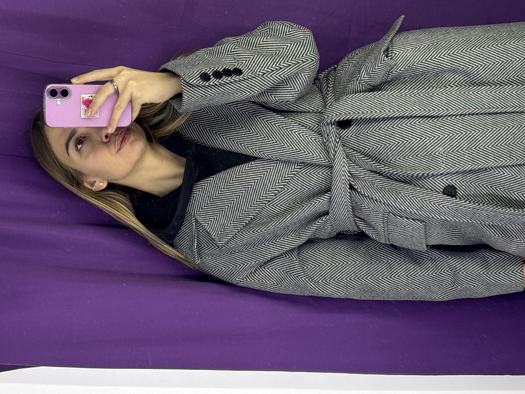 A woman lies on a purple surface, possibly a bed or a blanket, taking a selfie with a pink smartphone that partially covers her face. She is wearing a gray and white herringbone pattern coat. The bottom of the image shows a white surface. The location is not determinable from this image.