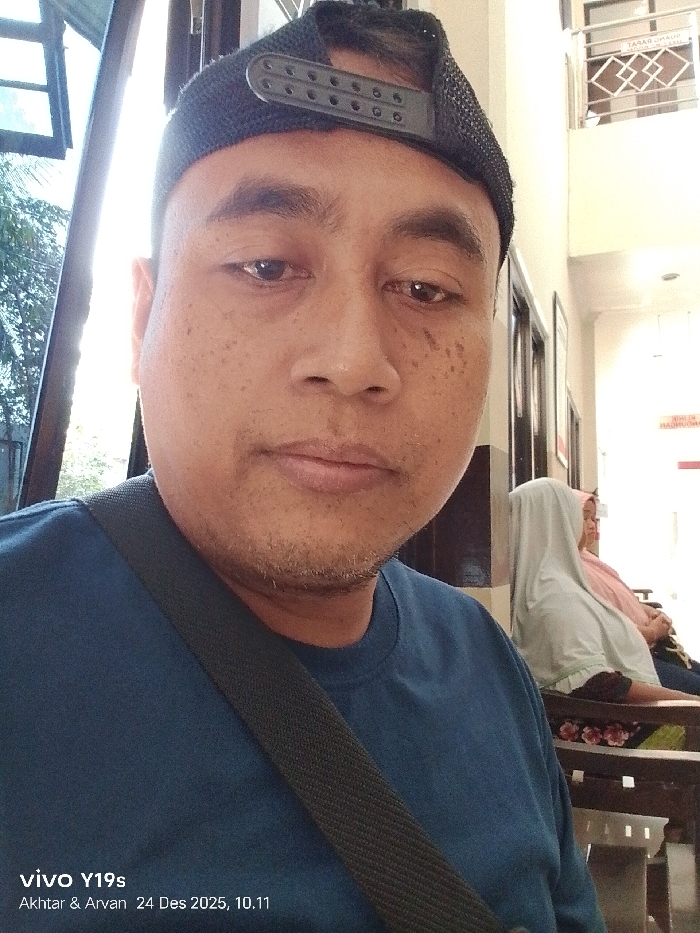 A man wearing a black hat and a dark blue shirt looks at the camera. He has a shoulder bag strap across his torso. Behind him are two women seated. One woman wears a white headscarf and the other wears a pink headscarf. The background features an interior space with windows and a sign in the distance. The image includes the text "vivo Y19s Akhtar & Arvan 24 Des 2025, 10.11" in the bottom left corner.