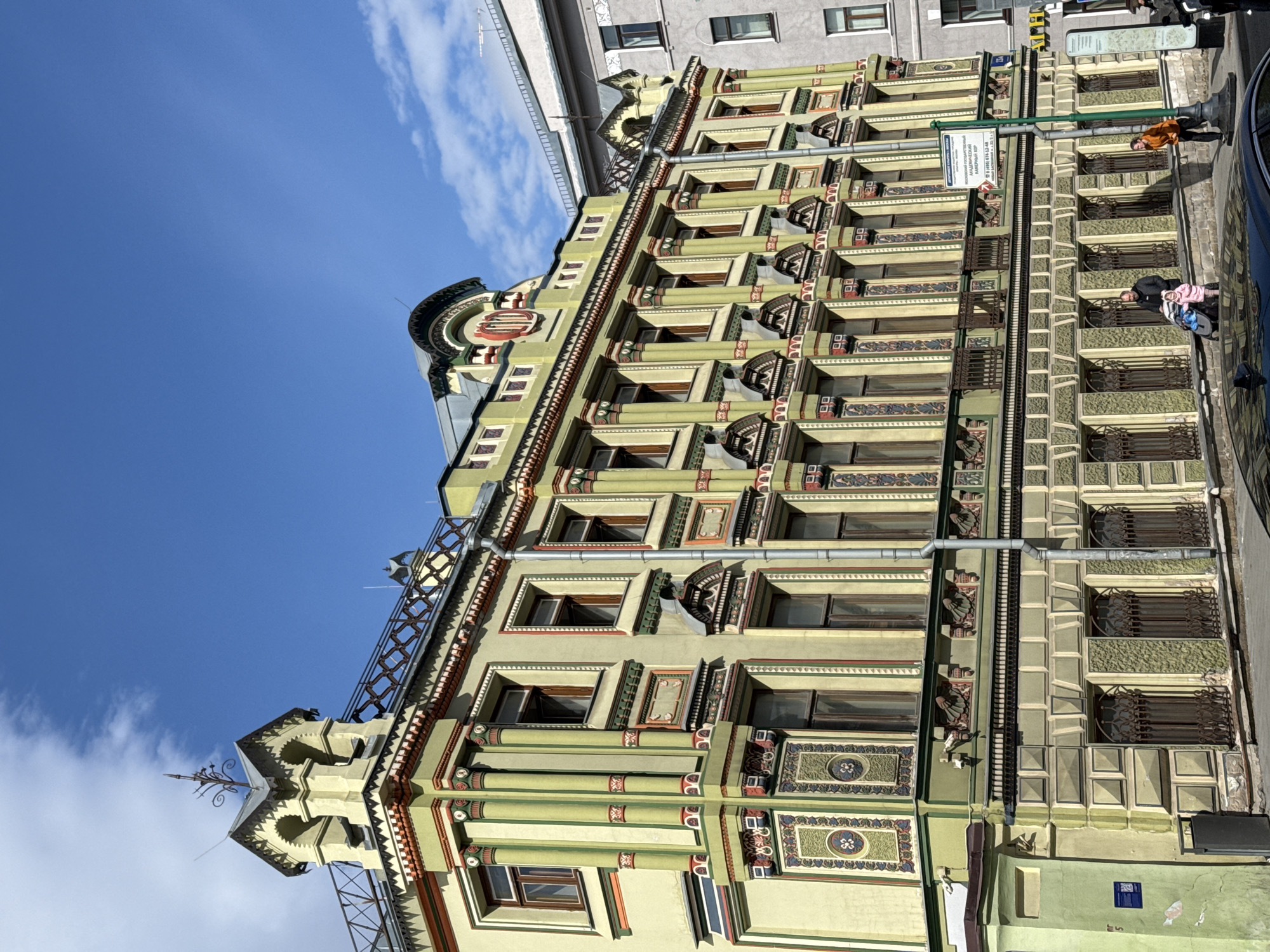 The image shows a detailed view of a historical building in Moscow, Russia. The structure is a light green color, adorned with intricate architectural details including decorative window frames and ornate columns. The building is likely of historical significance, based on its distinctive design and the presence of a street sign written in Russian. The sky above is bright blue with some wispy clouds. On the street level, there are a few pedestrians and a parked car.