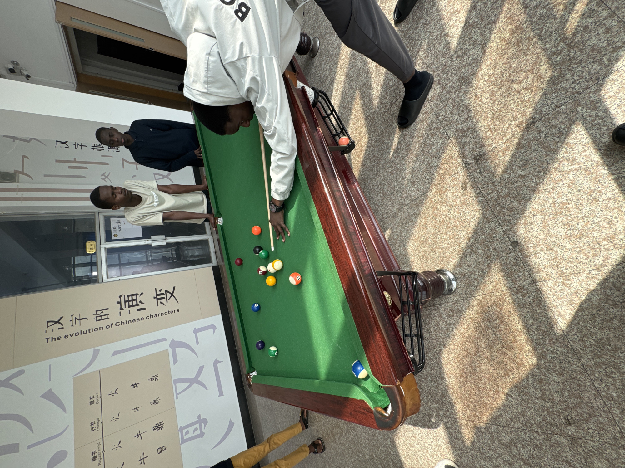 The image depicts an indoor scene where several young men are gathered around a pool table. A central figure, wearing a light-colored jacket with a large 'B' on the back, is actively playing pool, leaning over the green-felted table with a cue stick, focused on making a shot. Numerous colored pool balls are spread across the table, with one blue and white ball partially pocketed and an orange ball in the ball return rack. Two other men observe the game from the opposite side of the table: one in a dark blue polo shirt and another in a light t-shirt. The legs of additional individuals are visible, indicating more people are present.
The setting appears to be within an educational institution in Zhongtun, China, characterized by prominent wall displays featuring Chinese language and culture. A large poster on the wall reads "汉字的演变" (The evolution of Chinese characters) and "The evolution of Chinese characters," showcasing different script styles like "楷书" (Regular script) and "行书" (Lineage/Cursive script), along with examples such as "汉字根源" (Origin of Chinese characters). Other visible text includes "教室" (classroom) and "汉语言4班" (Chinese Language Class 4).
The floor is made of light-colored, speckled granite tiles, on which distinct, elongated shadows are cast by the pool table and unseen structures, suggesting bright natural light entering from an angled source, indicating it is daytime. Security cameras are mounted on a wall in the background. The scene portrays a casual, recreational activity taking place in an educational environment.
