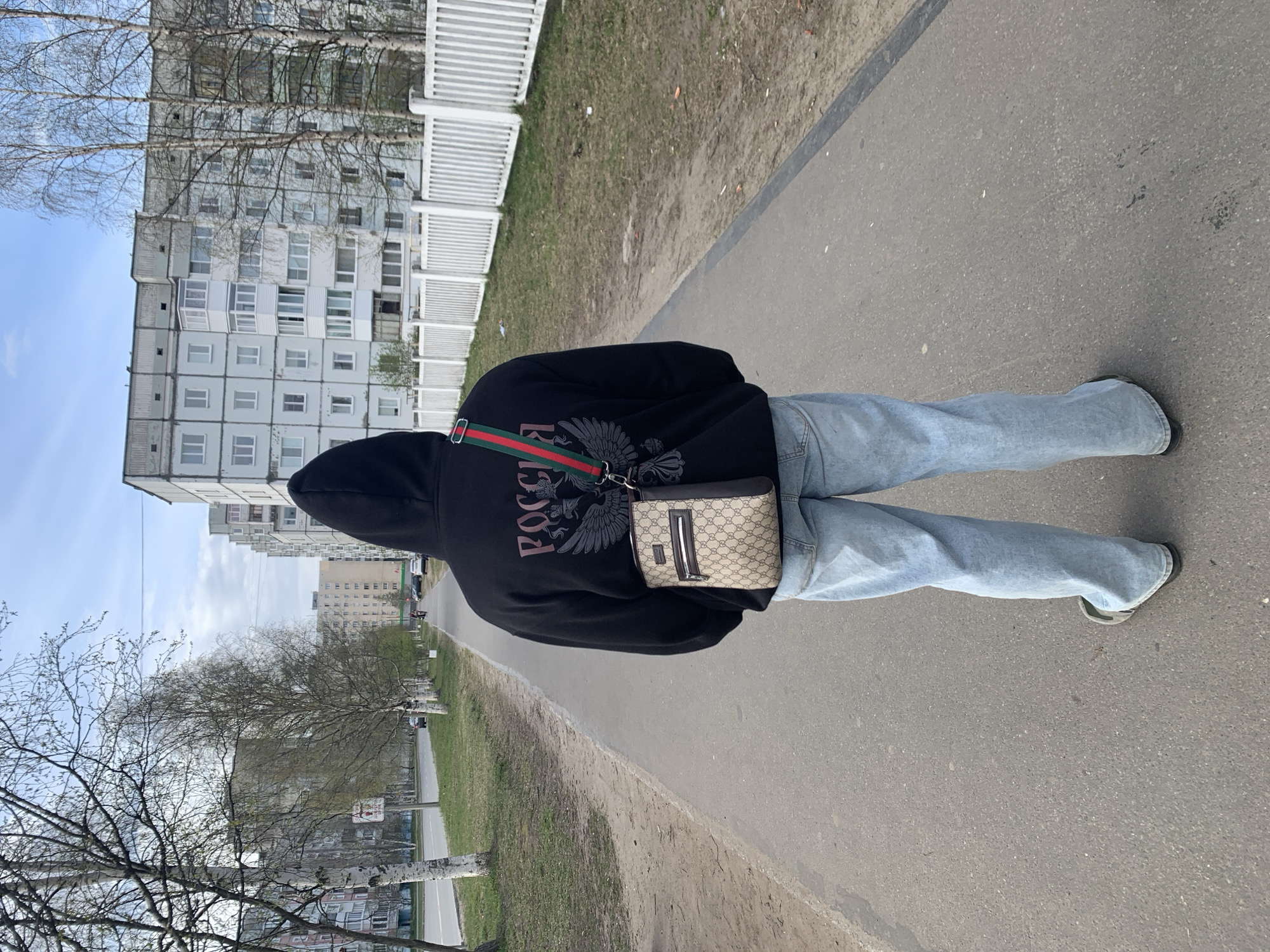 A person wearing a black hooded sweatshirt with Russian flag imagery and a small Gucci bag is standing on a concrete sidewalk. They are wearing light blue jeans and appear to be in motion, possibly walking away from the camera. The background includes a large, Soviet-era apartment building with many windows and a white fence. A grassy area borders the sidewalk on one side. Bare trees are present in the foreground. The scene appears to be in an urban environment. The location may be in Severodvinsk, Russia, but not enough information to confirm.