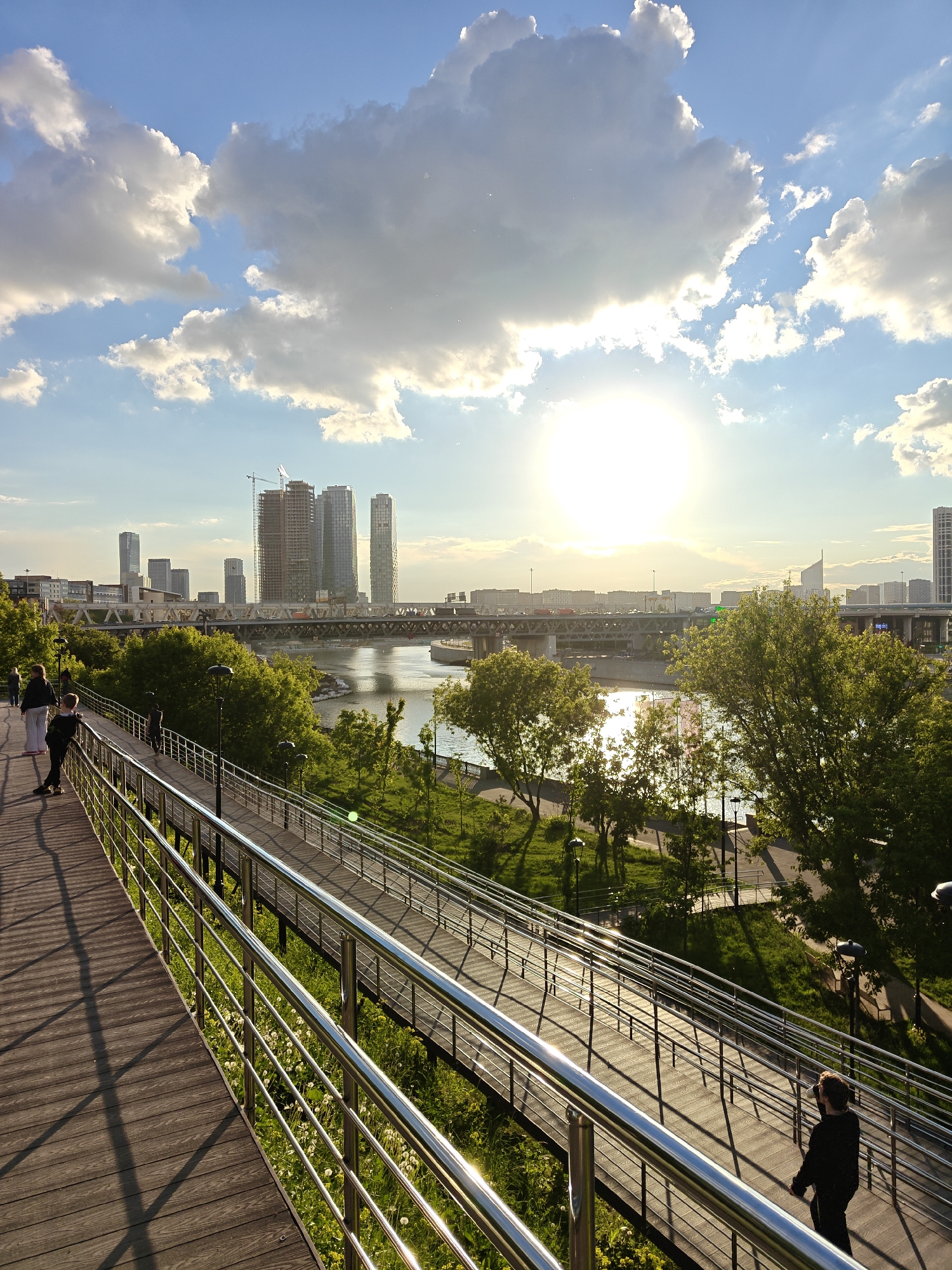 The image captures a sunny day scene in Moscow, Russia. In the distance, the Moscow International Business Center skyline is visible, with several skyscrapers, including one under construction with a prominent crane. A large bridge spans the river, carrying traffic.
In the foreground, a modern pedestrian walkway with stainless steel railings extends along the riverbank. Several people are present on the walkway, including a person wearing black on the right side of the image. Lush green trees and grass fill the space between the walkway and the river.
The sun shines brightly in the sky, partially obscured by clouds. The sky is blue with scattered white clouds. The image is well-lit, with the sun casting shadows on the walkway.