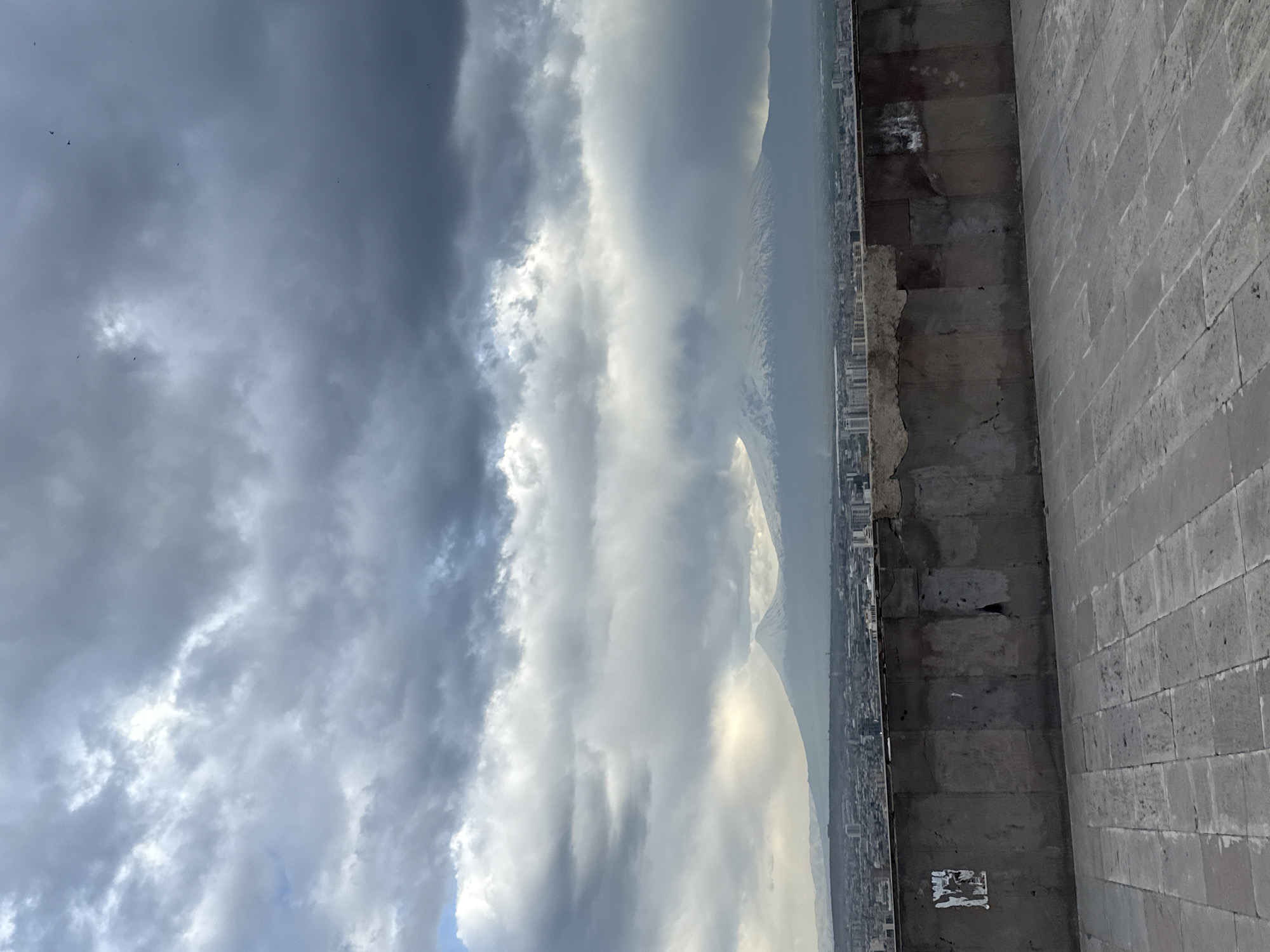 The image shows a view from a high vantage point in Yerevan, Armenia. A stone wall and brick walkway form the foreground, looking out over the city and a distant, snow-capped mountain range. The sky is filled with dramatic, layered clouds, ranging from dark gray to bright white, indicating potentially stormy weather. Small, dark objects resembling birds are visible in the upper left corner of the sky.