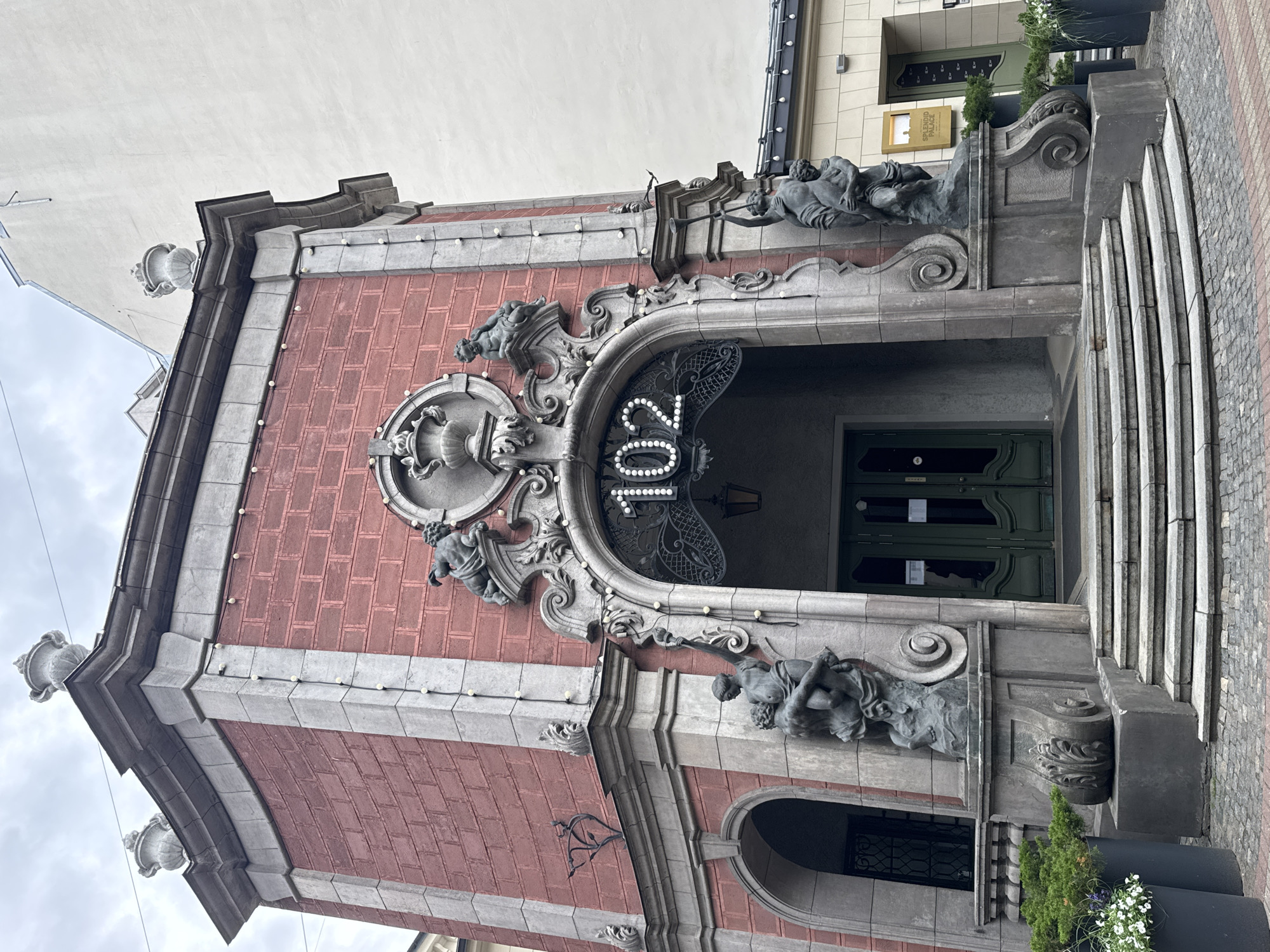 This is a medium shot, eye-level photograph of the exterior of a building at 102 Elizabetes iela in Riga, Latvia. The building has a red brick facade with ornate stone detailing. The entrance features a large arched doorway with a metal gate and the illuminated number "102" above it. Two large stone sculptures of figures are positioned on either side of the entrance, flanking a set of stone steps leading up to the door. There are also decorative urns on top of the building. The sky is overcast. The scene is devoid of people.
