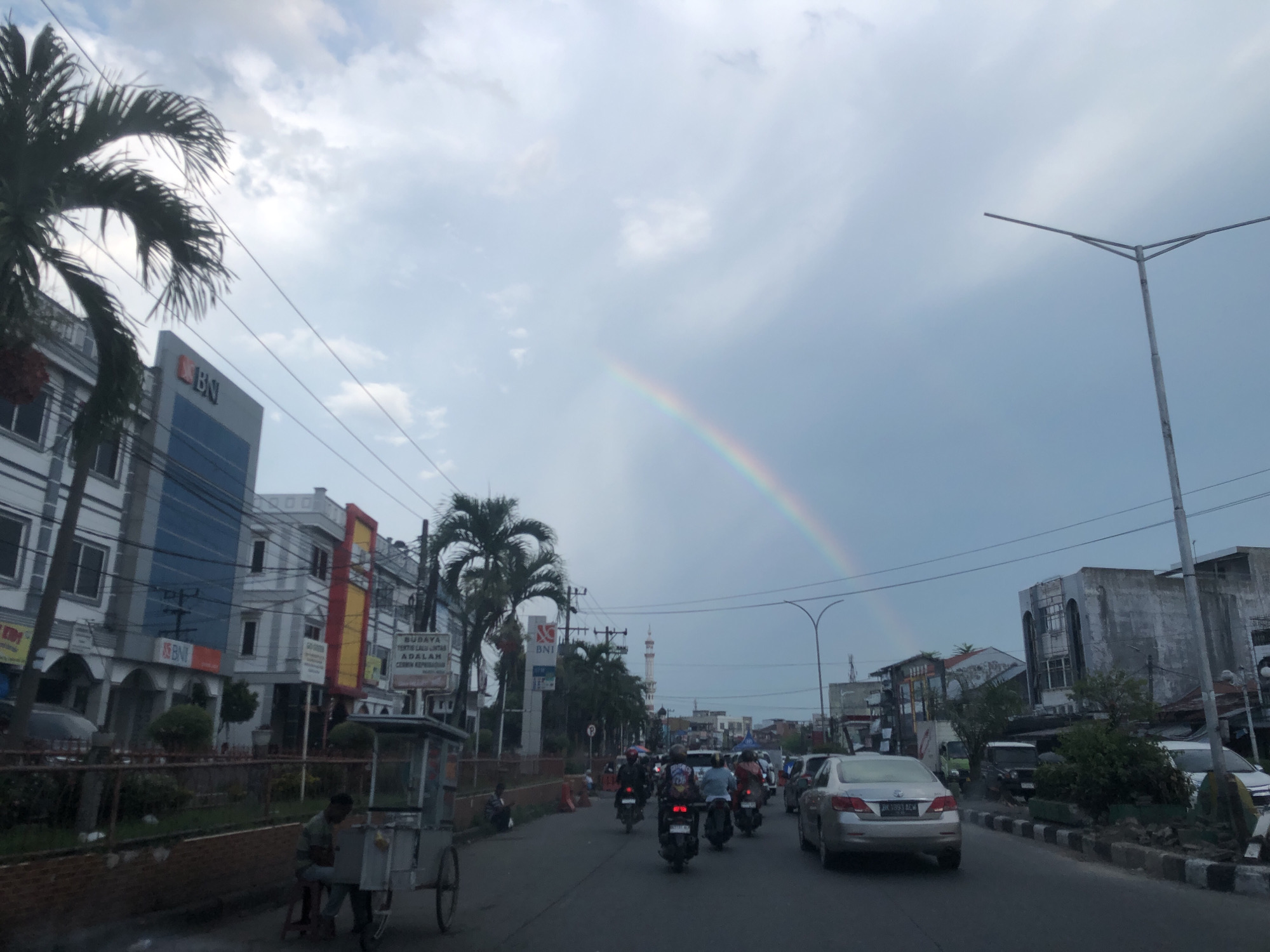 The image depicts a street scene in Medan, Indonesia, with a rainbow visible in the sky. In the foreground, a street vendor sits beside his cart on the left side of the road. A group of motorcycles and a silver sedan are traveling along the road. Buildings line both sides of the street; on the left, a BNI bank building is visible, and a mosque minaret is discernible in the distance. Palm trees are interspersed among the buildings. A street light stands on the right side of the road. The sky is overcast with the distinct rainbow stretching across the horizon.