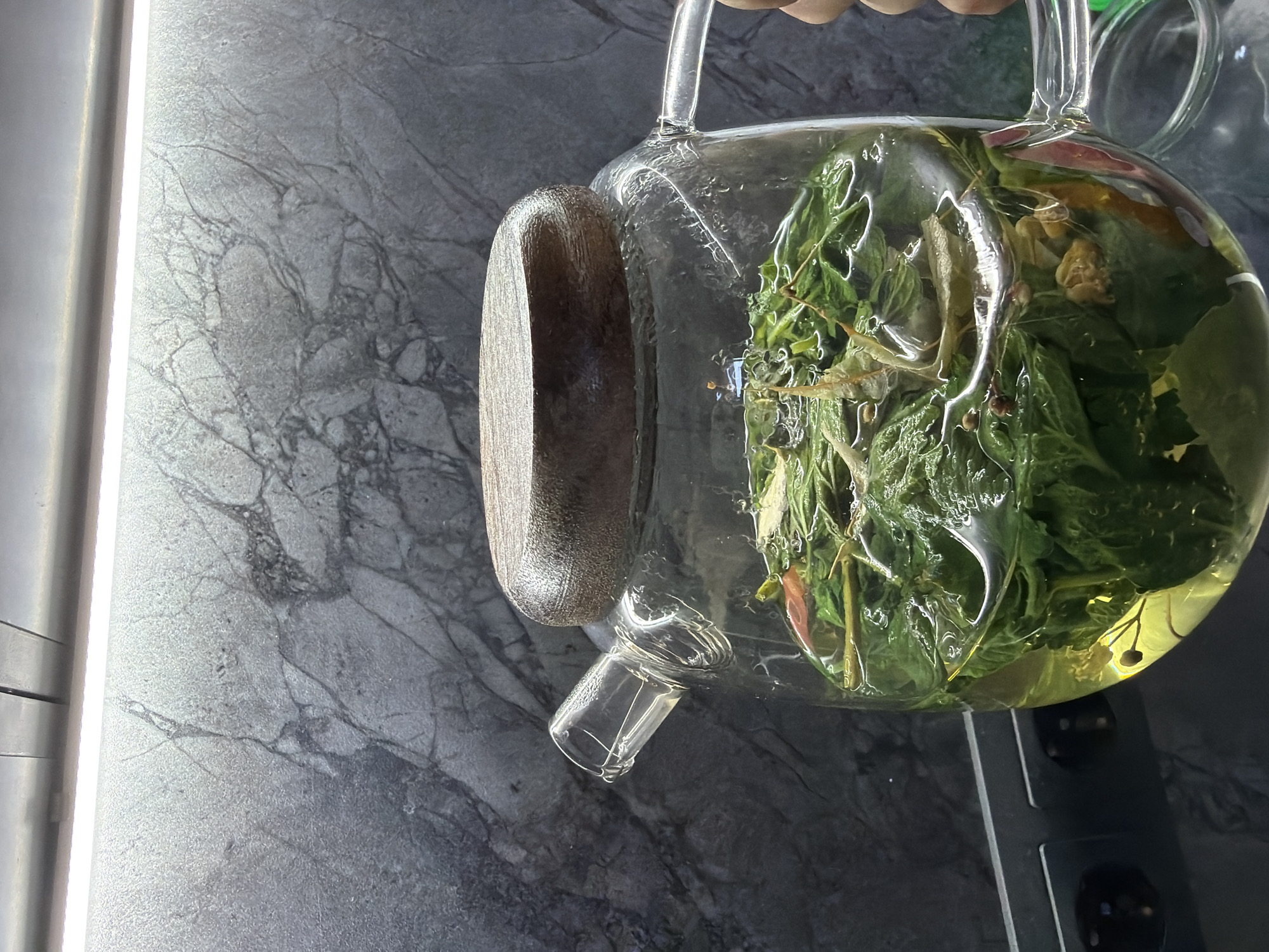 A glass teapot filled with green herbs and a yellow liquid is being held over a gray countertop. The countertop has a marbled pattern. The scene takes place in Chaikovsky, Russia.