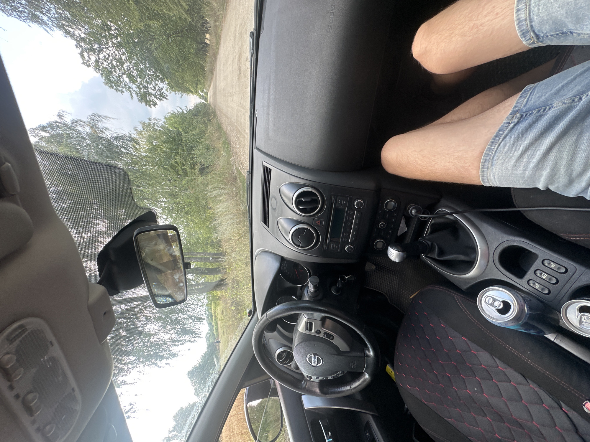 The image shows the interior of a Nissan car, looking forward through the windshield. The driver's side of the car is visible, including the steering wheel and dashboard. The gear stick is in view, with two open cans of soda next to it. The passenger's legs are visible in the foreground, wearing denim shorts. The car is on a dirt road lined with trees, suggesting a rural or semi-rural location. The sky is cloudy. The location in Tynna, Ukraine, is not explicitly depicted in the image, but the environment suggests a natural setting.