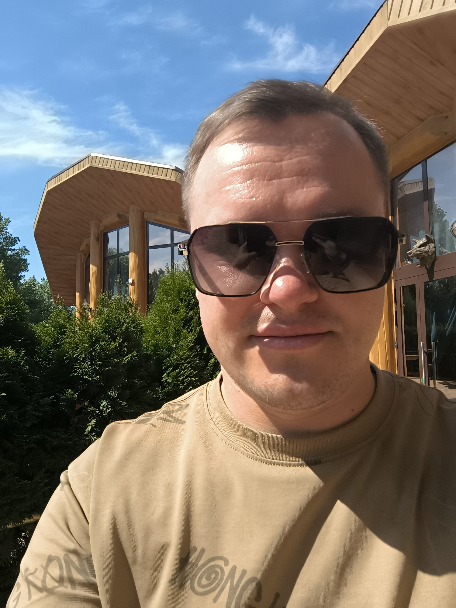 A man, wearing sunglasses and a light brown t-shirt with dark grey lettering, stands in front of a wooden building with large windows. Lush green bushes are visible in the foreground. The sky is blue with wispy white clouds. The building appears to be a lodge or a similar structure, possibly located in Malyy Baklaniy, Russia. The man is positioned slightly off-center to the right, with his face taking up a significant portion of the frame. The lighting suggests it is a sunny day.