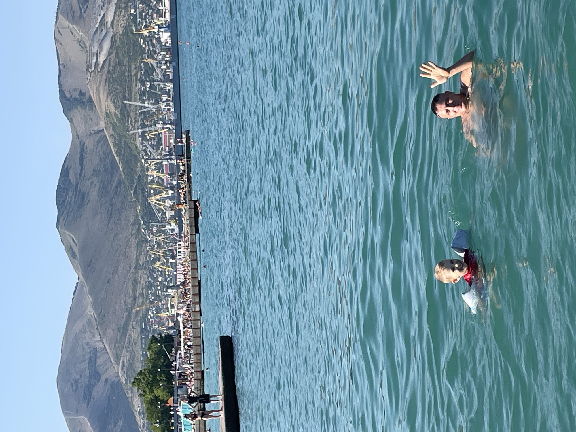 In the foreground, two individuals are swimming in clear, blue-green water. A young boy, wearing a red shirt and arm floaties, is smiling and looking towards the camera. To his right, a man is waving at the camera, with his upper body visible above the water. In the background, a pier extends into the water with several people standing on it. Beyond the pier, a city skyline is visible, characterized by numerous cranes and industrial structures, nestled at the base of a large, mountainous backdrop. The sky is clear and blue, indicating a sunny day. The scene appears to be a coastal area within the city of Sadovyi, Russia, likely a beach or harbor area given the presence of the pier and port facilities.