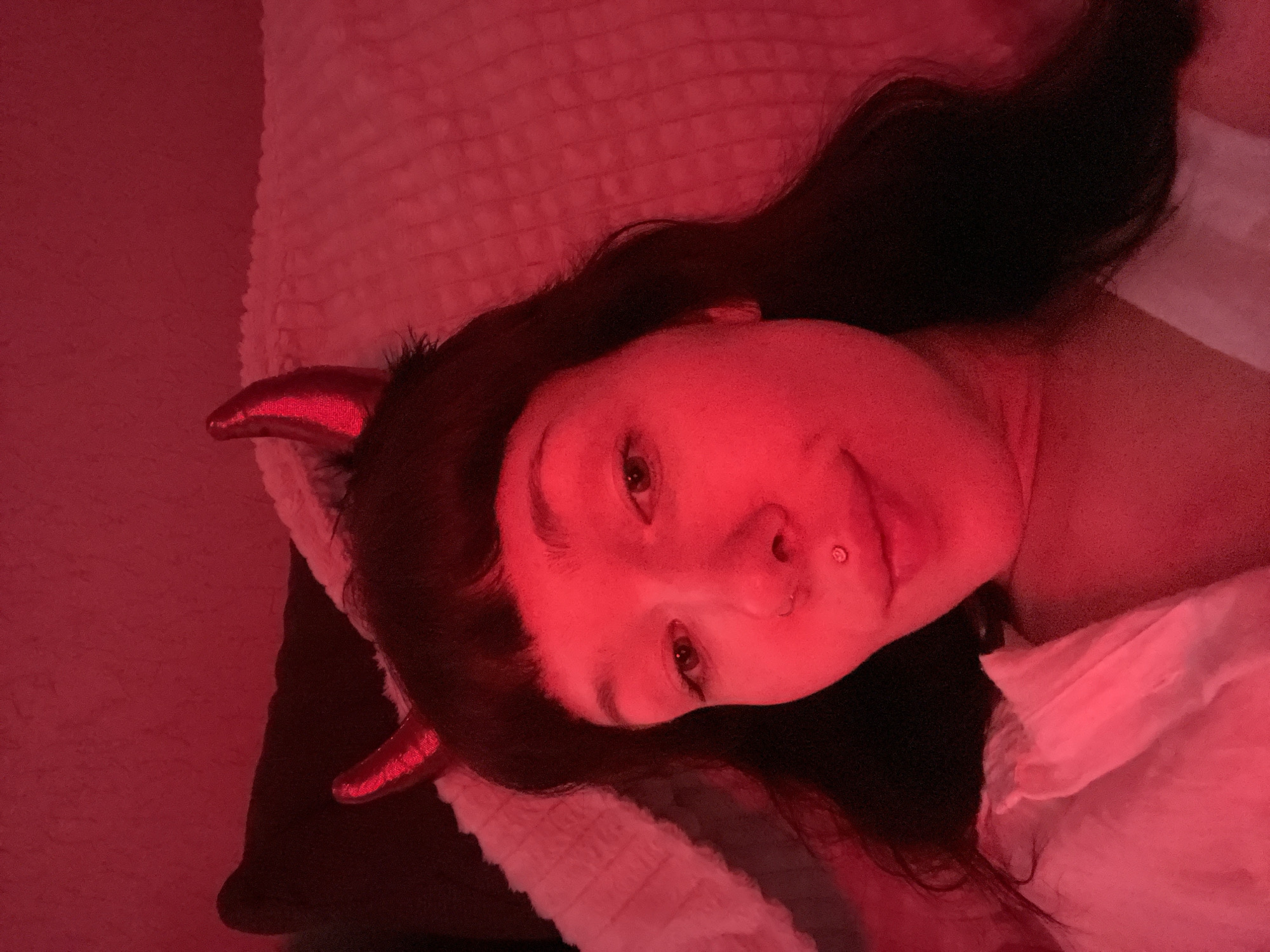 A woman with dark hair and red devil horns lays on a bed in a room illuminated by red light. She has fair skin, and a piercing in her nose. She is lying on a white patterned blanket, on what appears to be a brown pillow. The location is not apparent from the image.