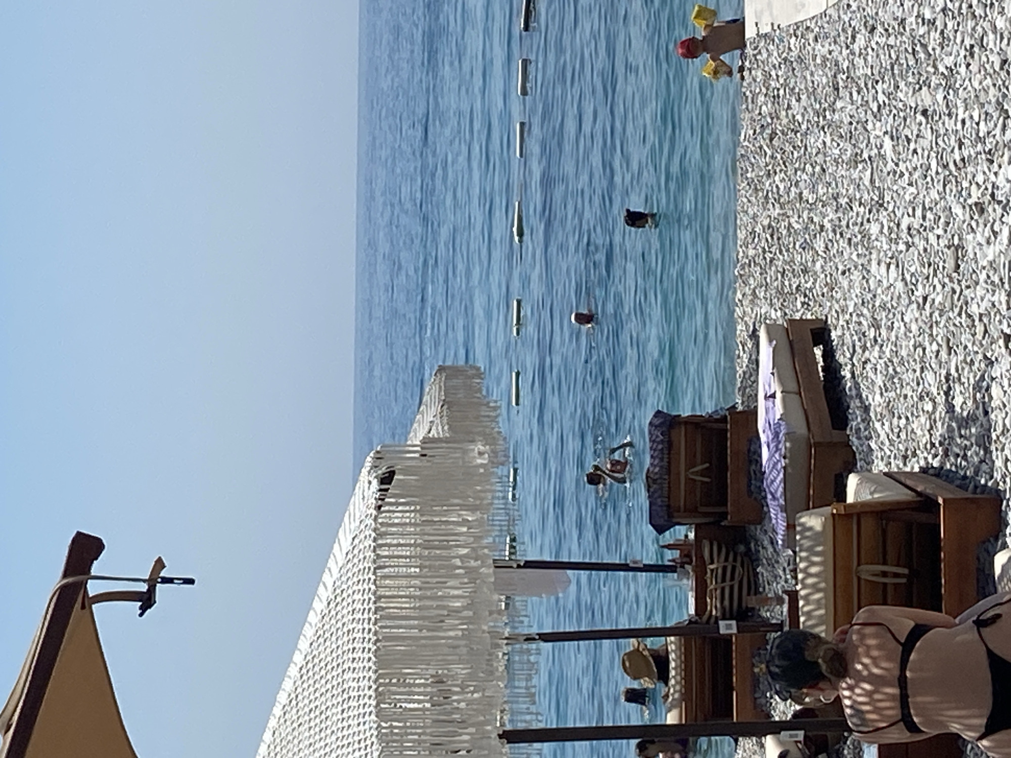 This image depicts a beach scene in Prijevor, Montenegro. The foreground shows a pebble beach with several lounge chairs and umbrellas, some occupied by people. In the middle ground, people are swimming and wading in the clear blue water. A rope barrier with buoys is visible in the water, marking a swimming area. The background consists of a clear blue sky meeting the horizon of the sea. The overall atmosphere is one of leisure and recreation on a sunny day.