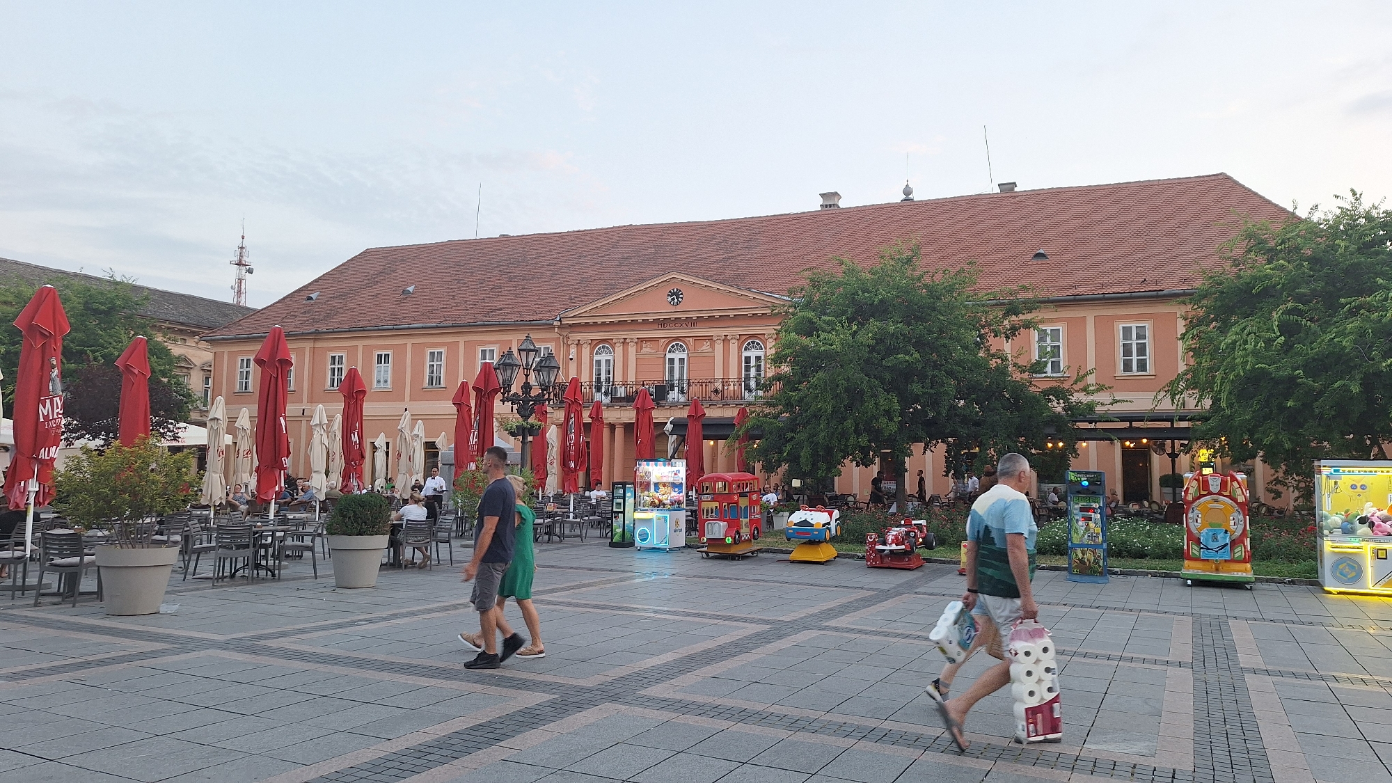 The image depicts a view of the central square in Sombor, Serbia. The square is paved with grey tiles and features several outdoor seating areas with tables and chairs, covered by numerous red and beige umbrellas. A large, orange building with a red tiled roof dominates the background, featuring arched windows and a central clock above a balcony. To the left of the main building, another building with a grey roof is visible, with a radio tower on top. Several trees are planted around the square, and there are also some children's amusement rides visible, including a red double-decker bus-themed ride, a yellow and blue ride, and a claw machine. In the foreground, two men are walking across the square. One man, on the left, is wearing a dark blue t-shirt and grey shorts, and the other man, on the right, is wearing a light blue and white striped t-shirt and white shorts, carrying several rolls of toilet paper. The sky is overcast.
