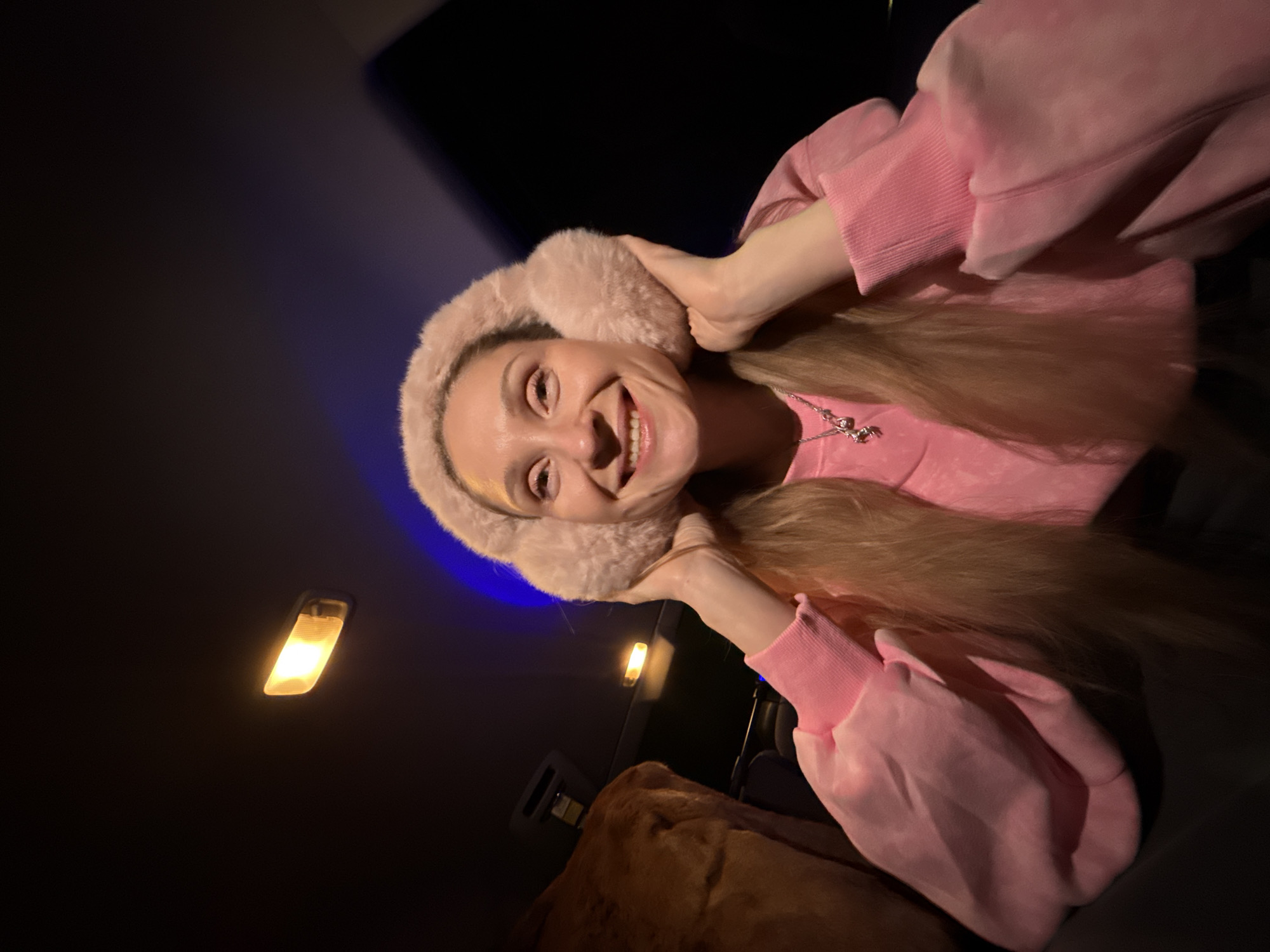 A woman is indoors, wearing a pink hooded top, a light pink fur-like hat, and a silver necklace. She is smiling and holding the sides of her hat. A car light is on in the background. Based on prior context, the location may be the city of Chaikovsky, Russia.