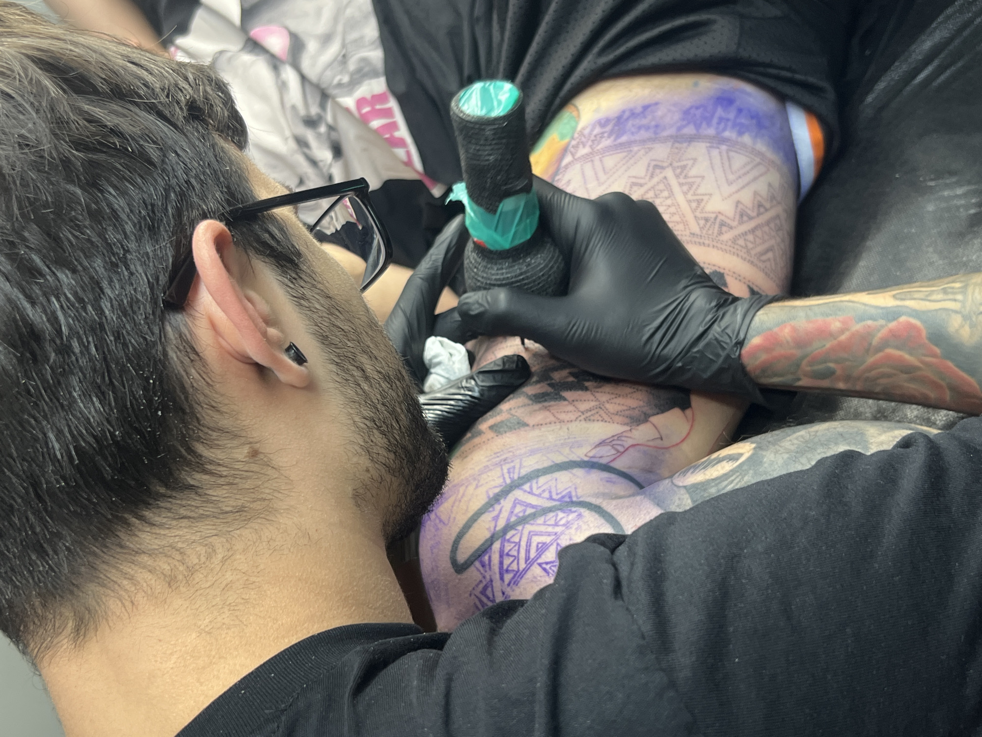 A person with short dark hair and a beard, wearing glasses and a black t-shirt, is seen from the side, tattooing someone. The person being tattooed is lying down and has a large geometric design in purple ink on their leg. The tattoo artist is wearing black gloves and holding a tattoo machine with a green-wrapped grip. The surrounding environment appears to be a tattoo studio. There is no definitive visual information to identify the location as Sofia, Bulgaria, or any other city.