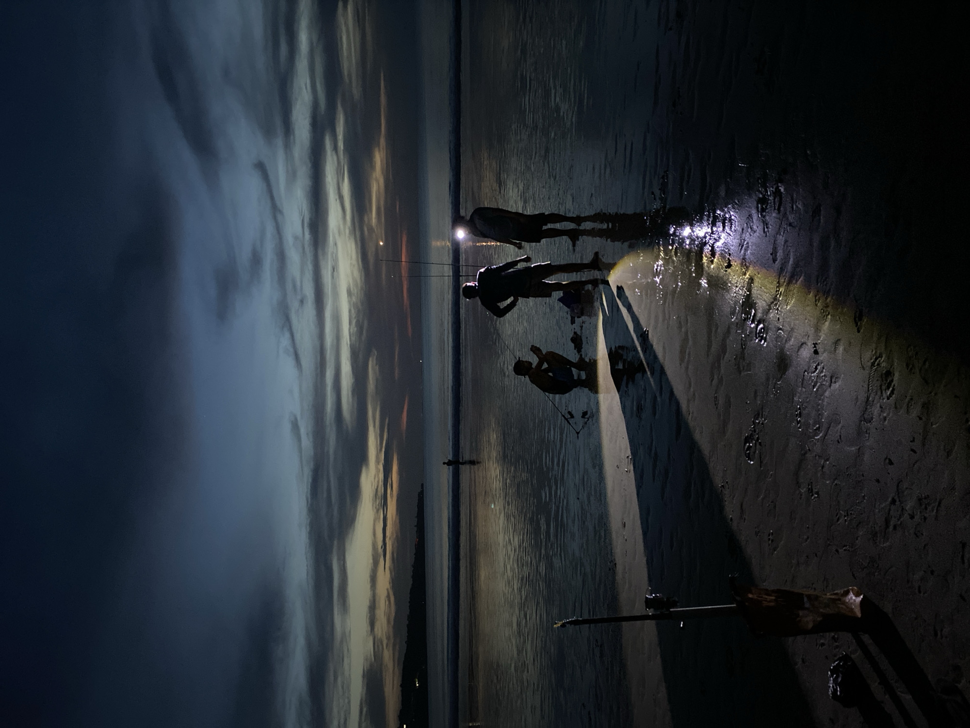 The image captures a night scene on a beach in Kuta Selatan, Indonesia. Three silhouetted figures are visible, illuminated by a strong light source that appears to be a flashlight. One figure is standing, another is crouching, and a third is standing behind the other two. The sand appears wet and reflects the light, creating a glossy surface. In the background, the sky shows a gradient from dark blue clouds to a lighter, sunset-like horizon. There are also fishing rods.