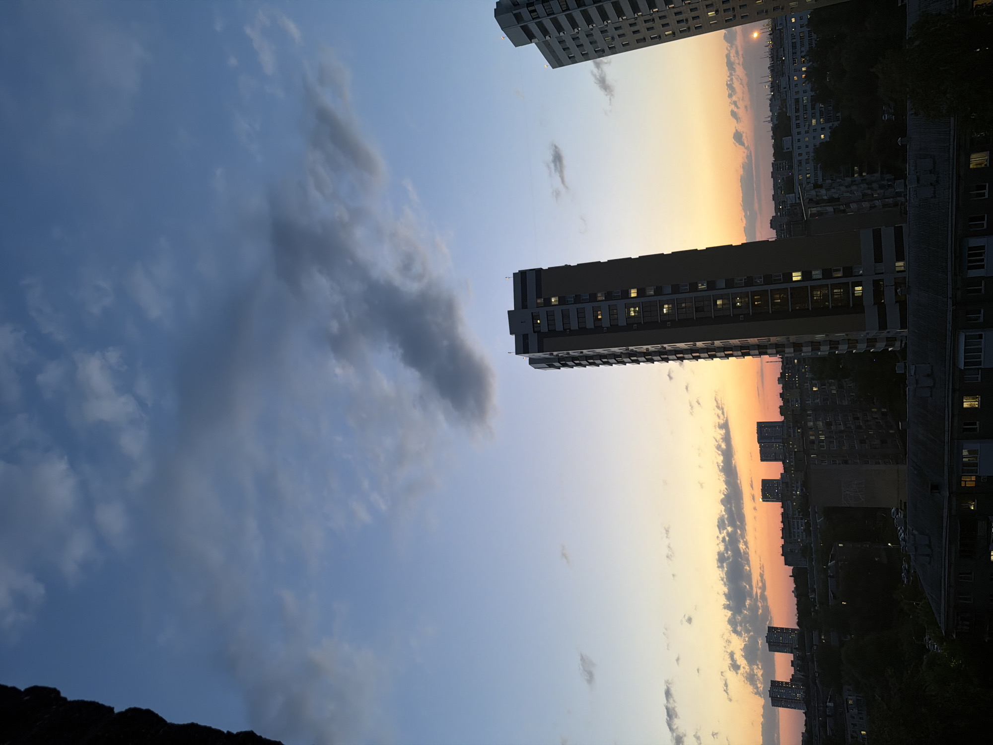 The image depicts a cityscape at sunset. Tall buildings are visible on either side of the frame, with a sky transitioning from blue to orange in the center. Some clouds are present in the sky. There are lights visible in some of the buildings. The location is in the city of Unknown, Unknown.