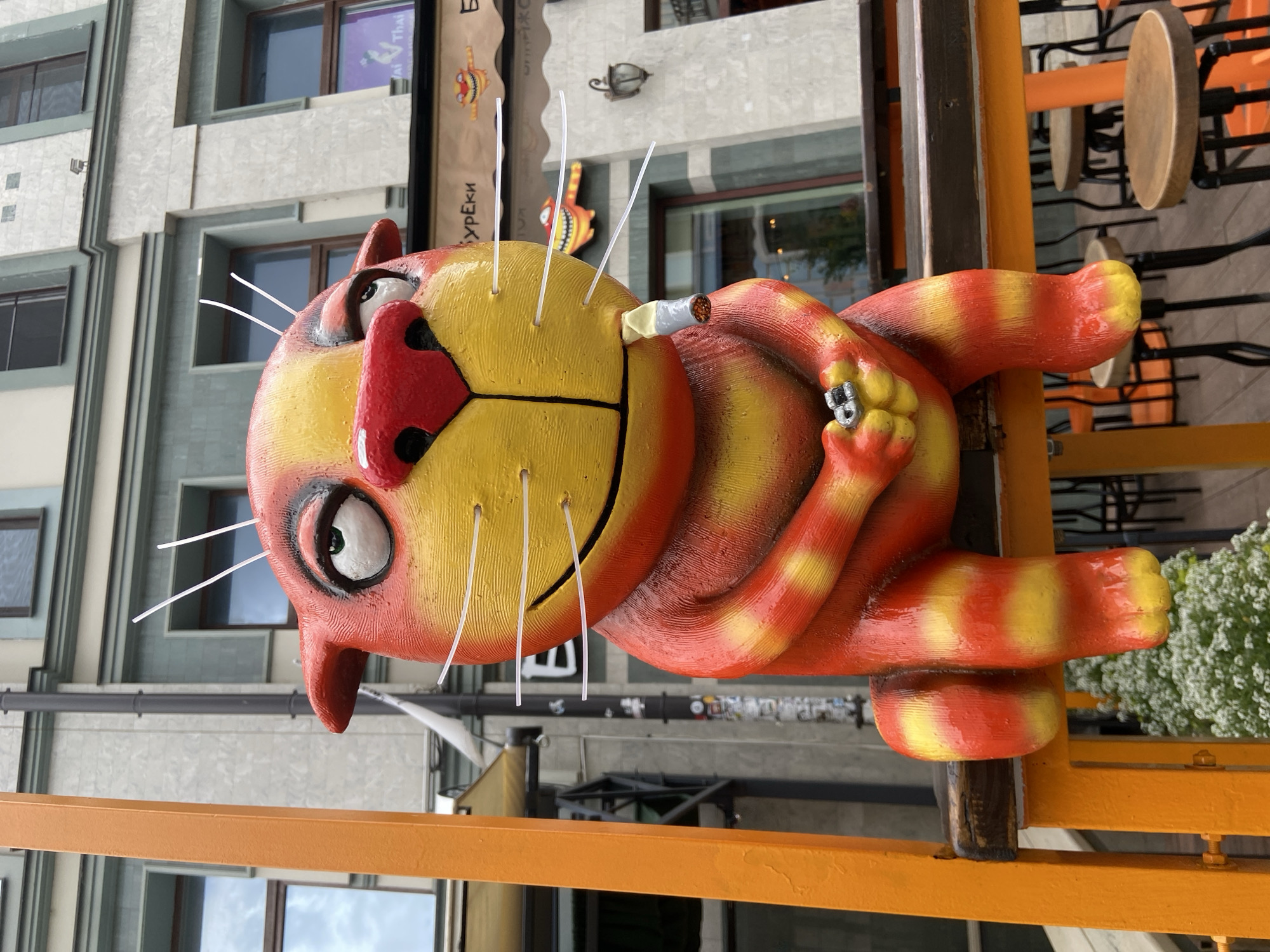 A street-level shot shows a statue of a cat with orange and yellow stripes that is smoking a cigarette. The cat is supported by an orange rail. The cat has a red nose, white whiskers, and is smiling. The statue is located in Kazan, Russia. Behind the statue is a building with windows and signs.