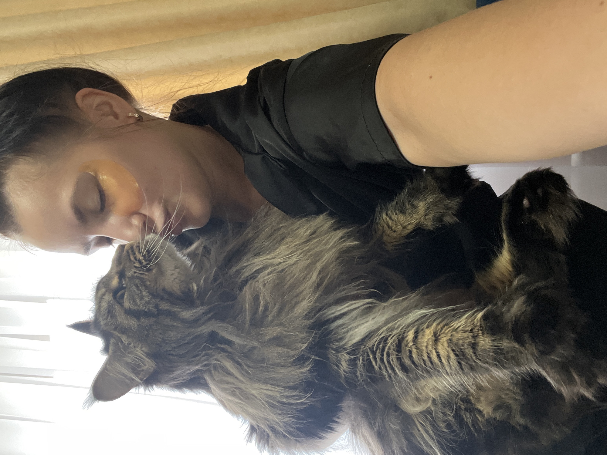 A woman with dark hair pulled back is holding a large, long-haired tabby cat. She is wearing a black top and has gold under-eye patches. The cat is nuzzling her cheek. The background consists of sheer white curtains and yellow drapes. The image appears to be taken indoors in what could be a residential setting in Марковский, Russia.