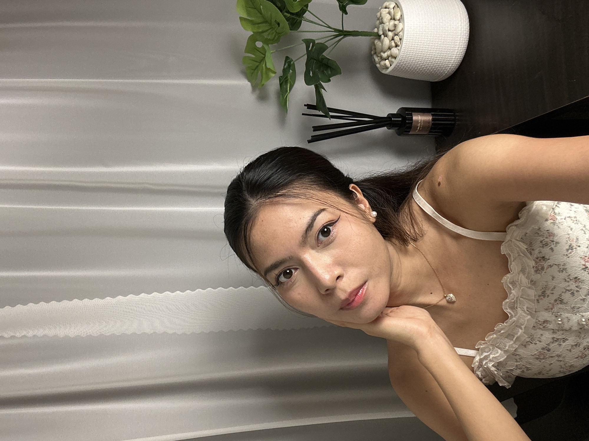 In this medium shot, a young woman is captured from the shoulders up, resting her chin on her hand. She is wearing a white lace top with ruffles and a delicate necklace with a heart-shaped pendant. Her hair is pulled back, and she has makeup on, including eyeliner and lipstick. She has a mole on her collarbone.
The background features a light gray, textured surface with horizontal lines, suggesting a curtain or blinds. To the upper right, a white textured pot holds a faux plant with green leaves and small white stones. Next to it is a black reed diffuser with a label that is too blurry to read. The lighting appears to be artificial, with a soft glow that highlights the woman's face and shoulders. The overall mood is relaxed and intimate, possibly a selfie taken indoors. The woman's expression is calm, with her gaze directed slightly away from the camera. The scene provides no direct clues about the time of day or weather.
