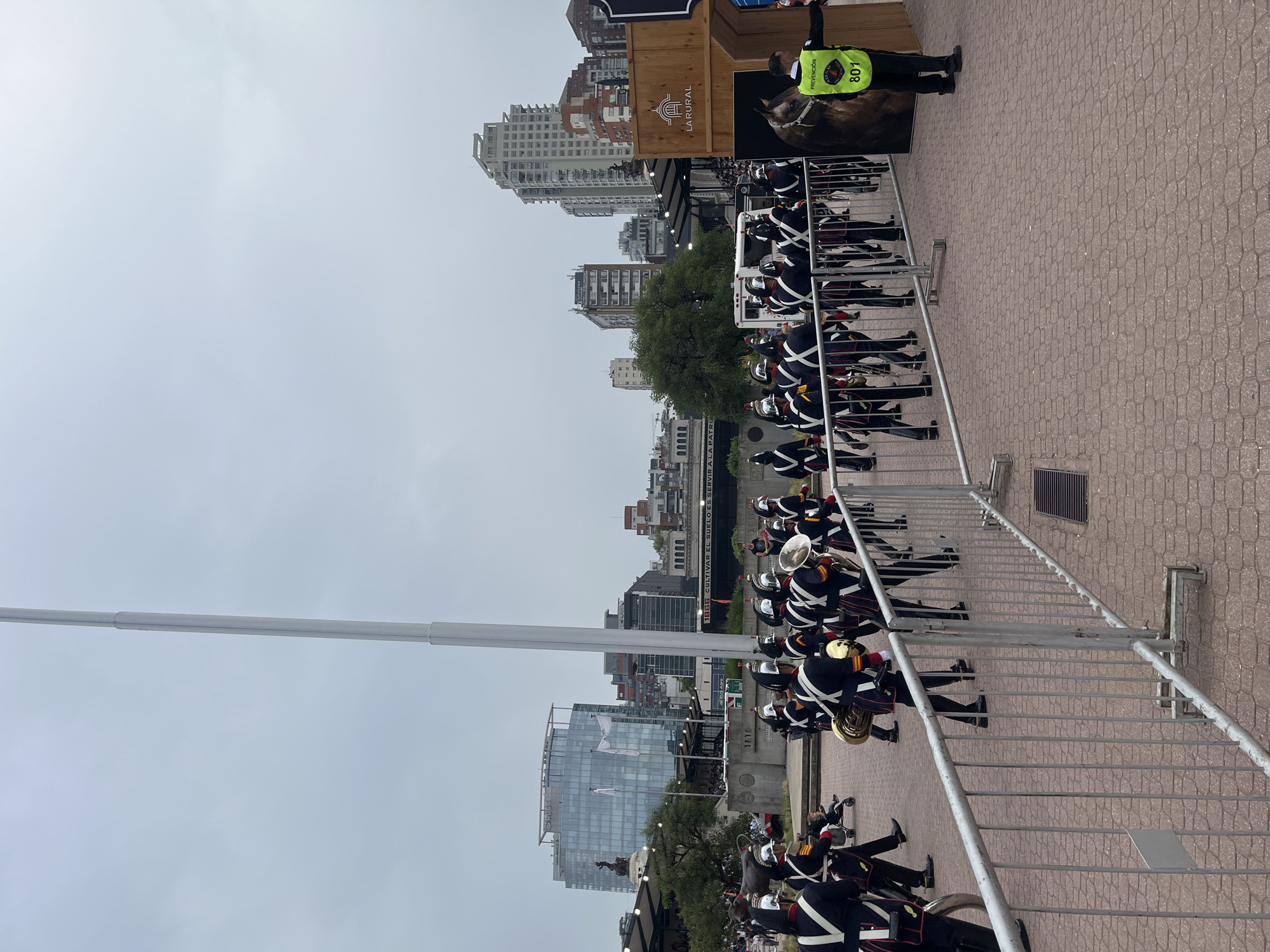 The image depicts an outdoor ceremonial event occurring on a wide, reddish-brown paver brick pathway at La Rural Exhibition Center in Palermo, Buenos Aires, Argentina. The sky is overcast.
In the mid-ground, a marching band, comprising approximately 15-20 individuals, advances from left to right. The band members wear dark blue or black uniforms with white cross belts, red trim, gold accents, and silver helmets adorned with plumes. Many are carrying brass instruments, including tubas, trombones, and possibly trumpets. Ahead of the marching band, grey metal barriers delineate the path.
To the far right, a security guard in a neon yellow-green vest, marked with "PREVENCIÓN" and "801", stands near a tall, wooden structure displaying the "LA RURAL" logo and a partial image of a horse's head. Behind the marching band, spectators are visible in tiered seating. A prominent, tall, white cylindrical flagpole stands to the left of the wooden structure. Further back, a concrete wall bears the inscription "1866 CULTIVAR EL SUELO ES SERVIR A LA PATRIA".
The background reveals a dense urban landscape featuring a mix of modern glass office buildings, one of which displays a "PLUS" logo, and various multi-story residential and commercial structures typical of Buenos Aires. To the far left, a dark statue, possibly depicting a bull or cow, is partially visible.